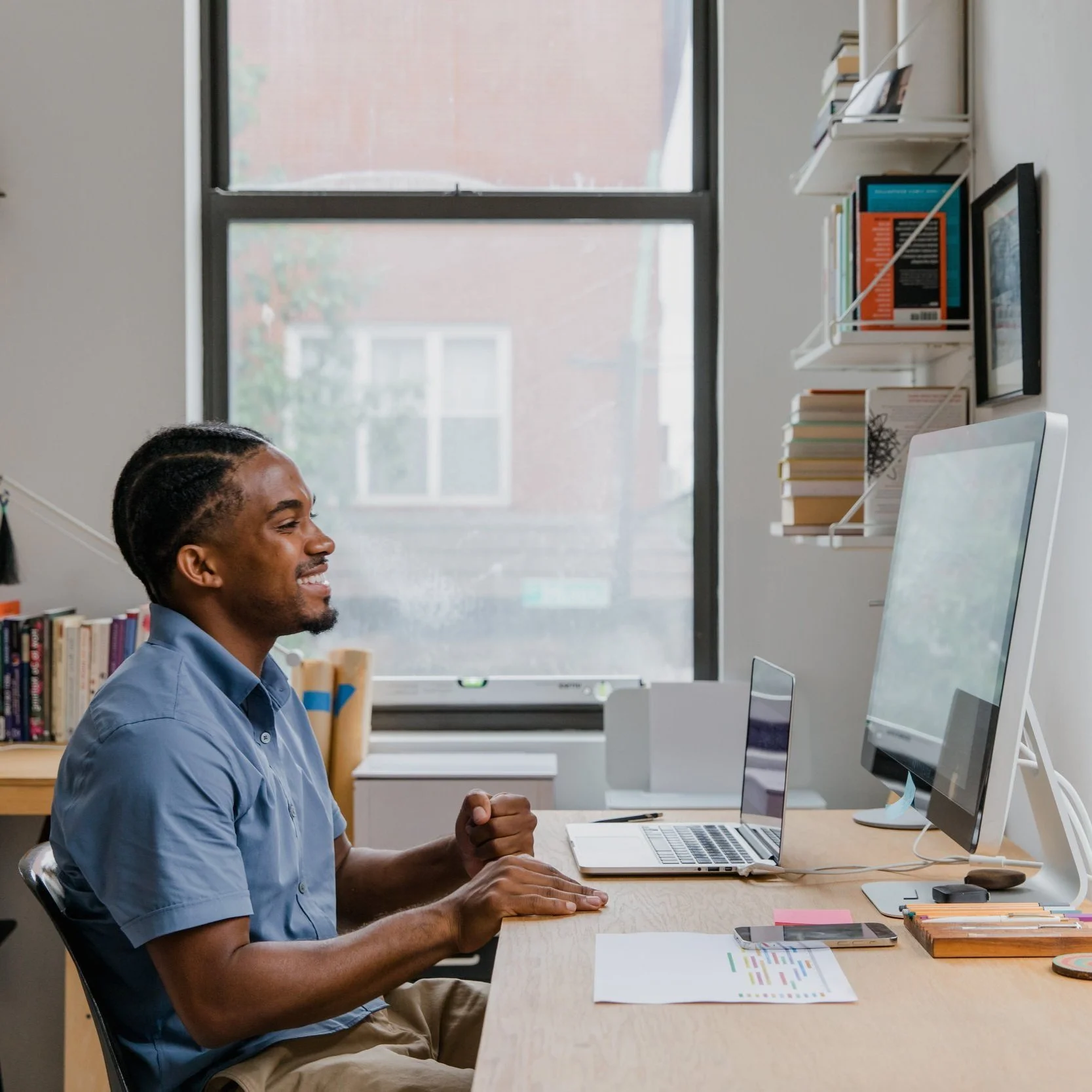 A man sitting at a desk in front of a computer, smiling with a fist clenched in celebration, in a well-lit room with a window, books, and office supplies.