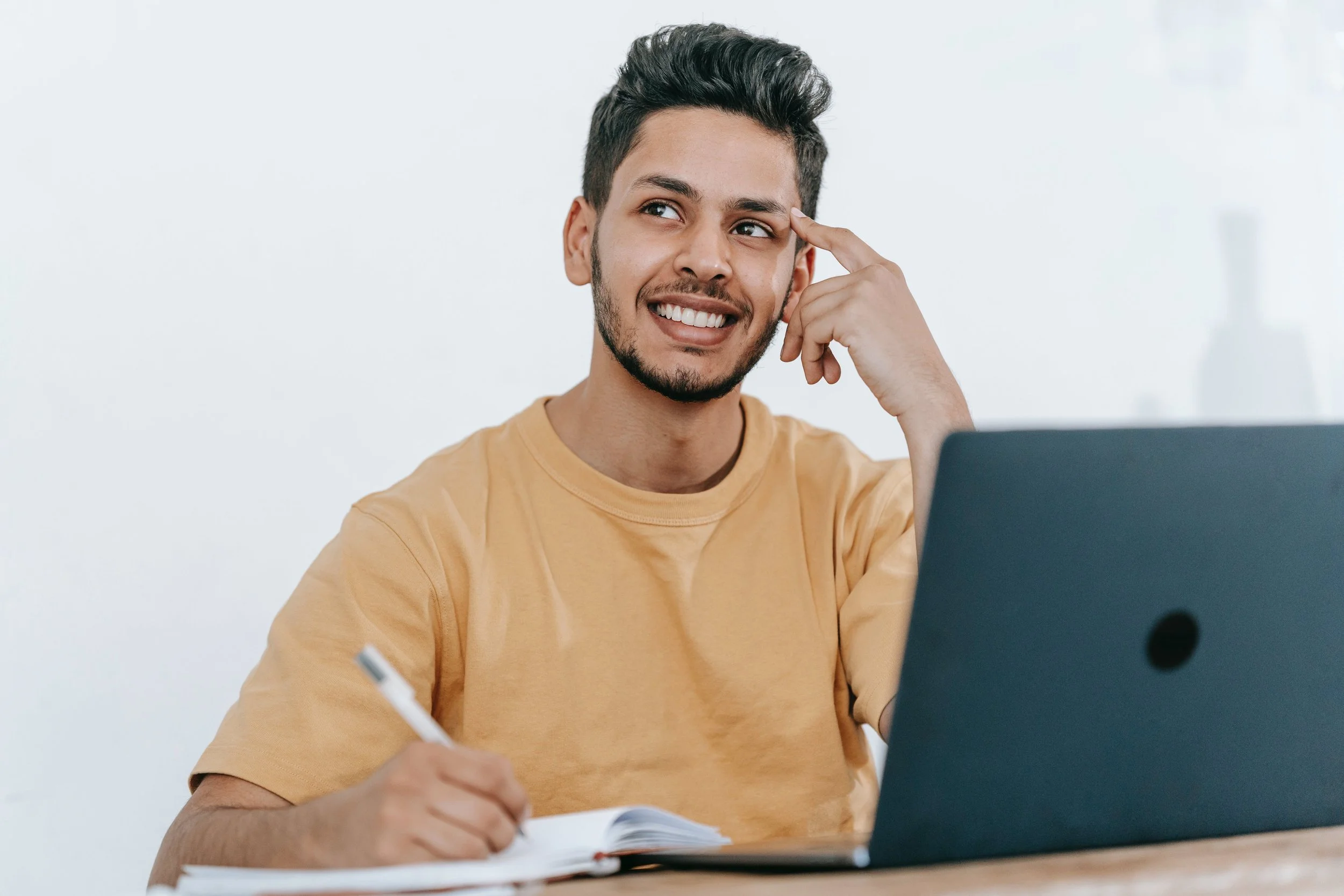 A young man with short dark hair and a beard, wearing a yellow t-shirt, sitting at a desk with a laptop and notebook. He is smiling and appears to be thinking, with one hand holding a pen over the notebook and the other touching his head.