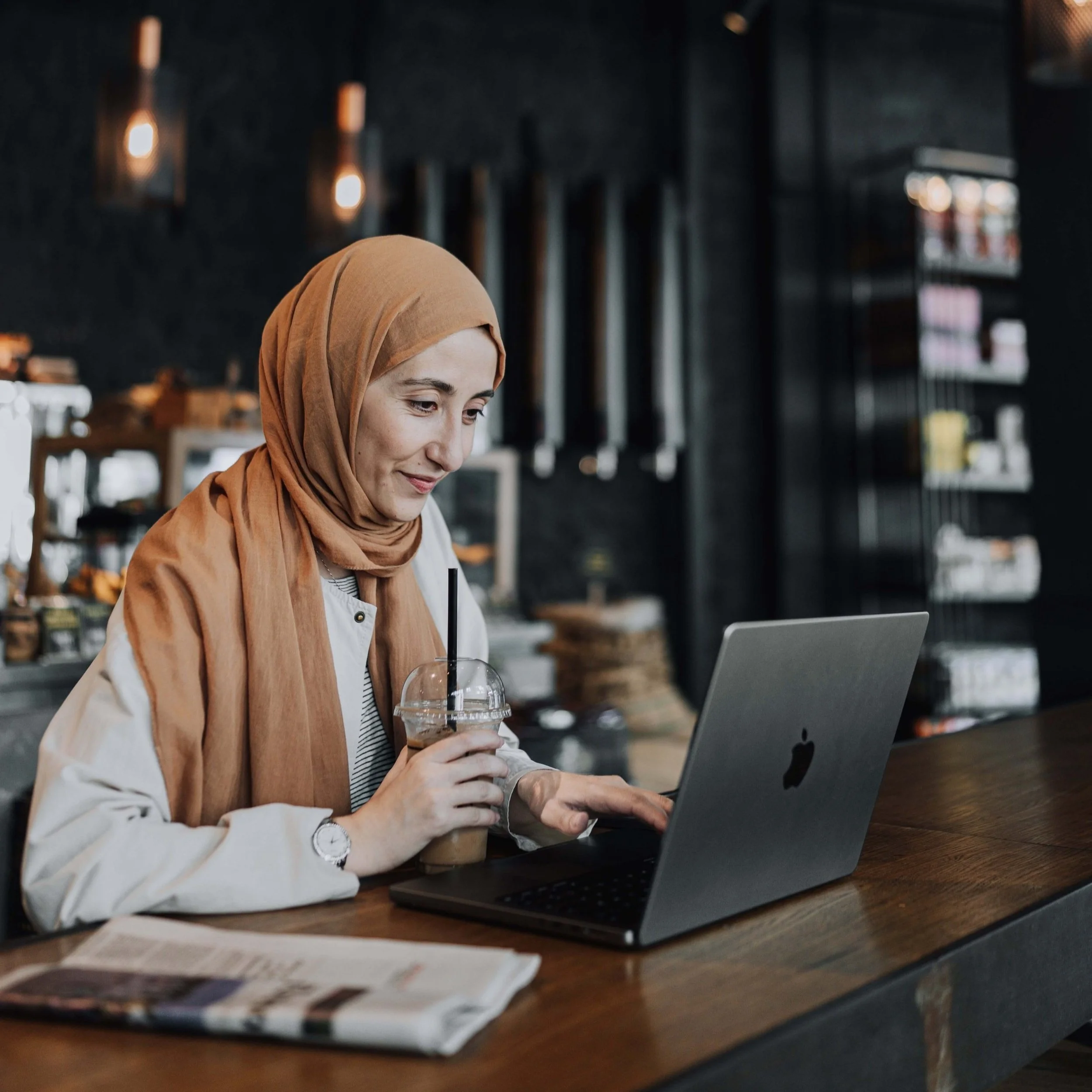 A woman wearing a tan hijab working on a laptop at a coffee shop, holding an iced coffee and reading a newspaper.