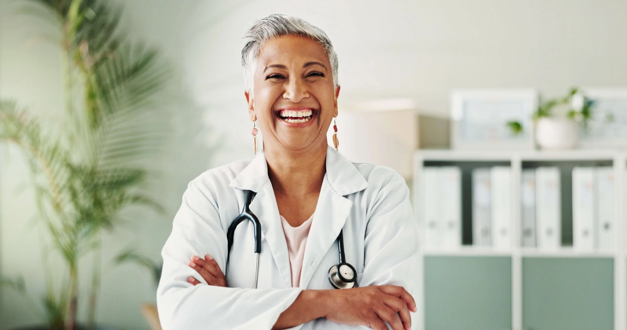 Smiling female doctor with short gray hair, white coat, and stethoscope, crossing arms in a medical office with green plants and shelves in the background.