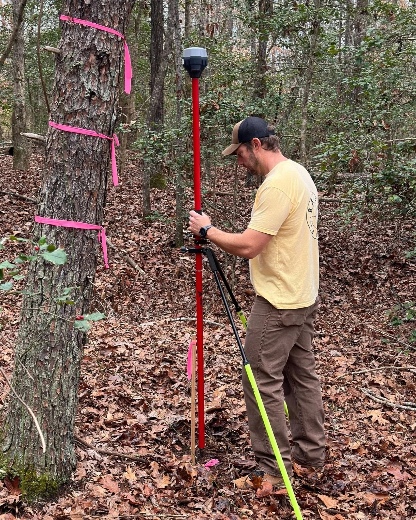 A man in a yellow shirt and brown pants using surveying equipment in a wooded area near Gastonia NC.