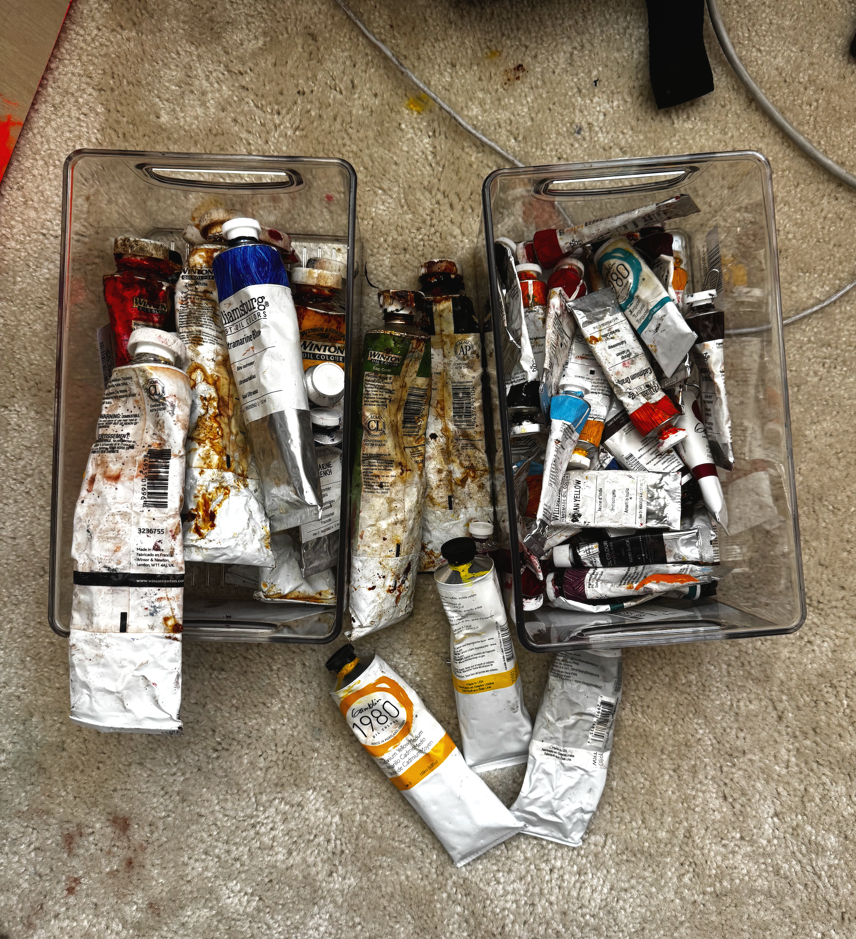 boxes of oil paint tubes from gamblin and williamsburg paints including colors like ultramarine blue, titanium white and cadmium yellow sitting on the floor of liz lancaster's art studio