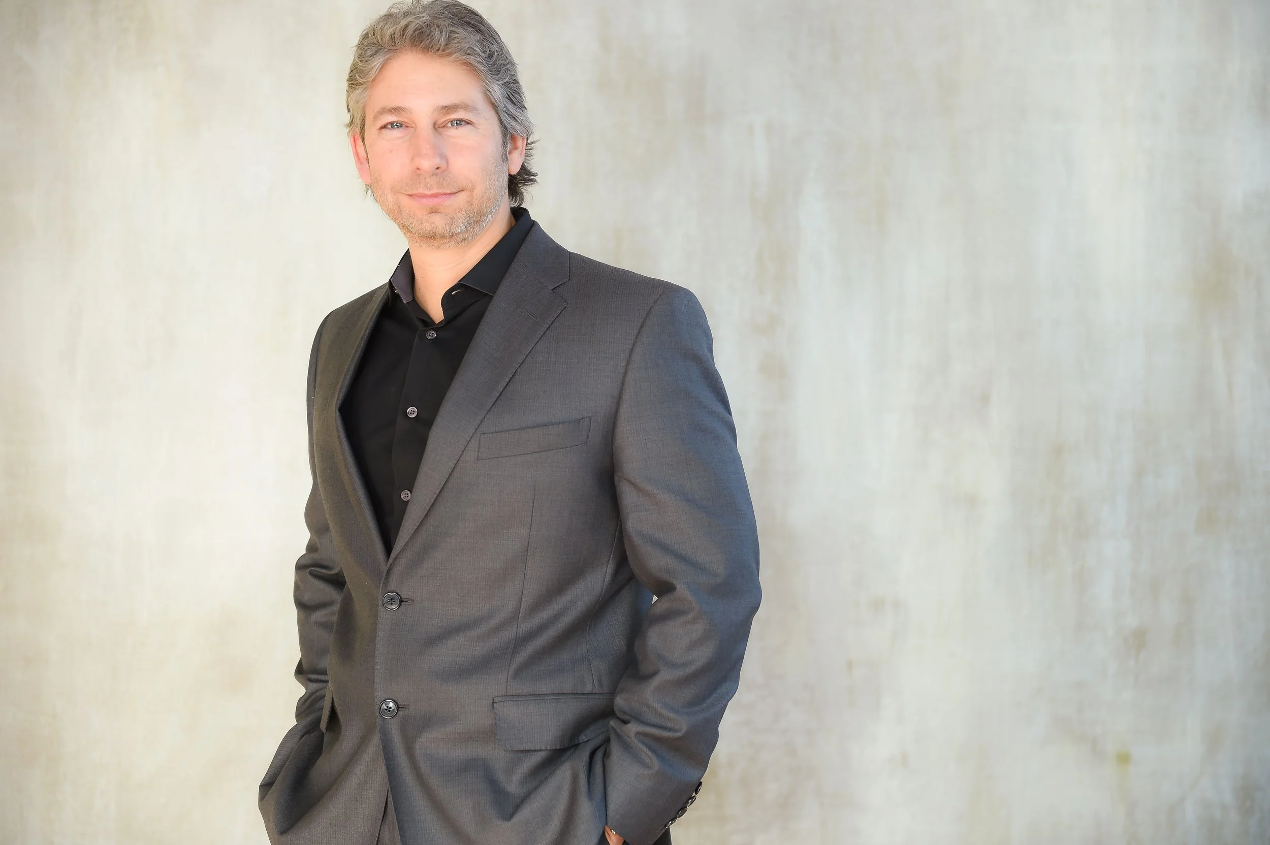 Andrew Goldwasser, a confident middle-aged lawyer man at Ciano & Goldwasser, in Beachwood, Ohio with gray hair and a beard, dressed in a gray suit and black shirt, standing against a light, textured background.
