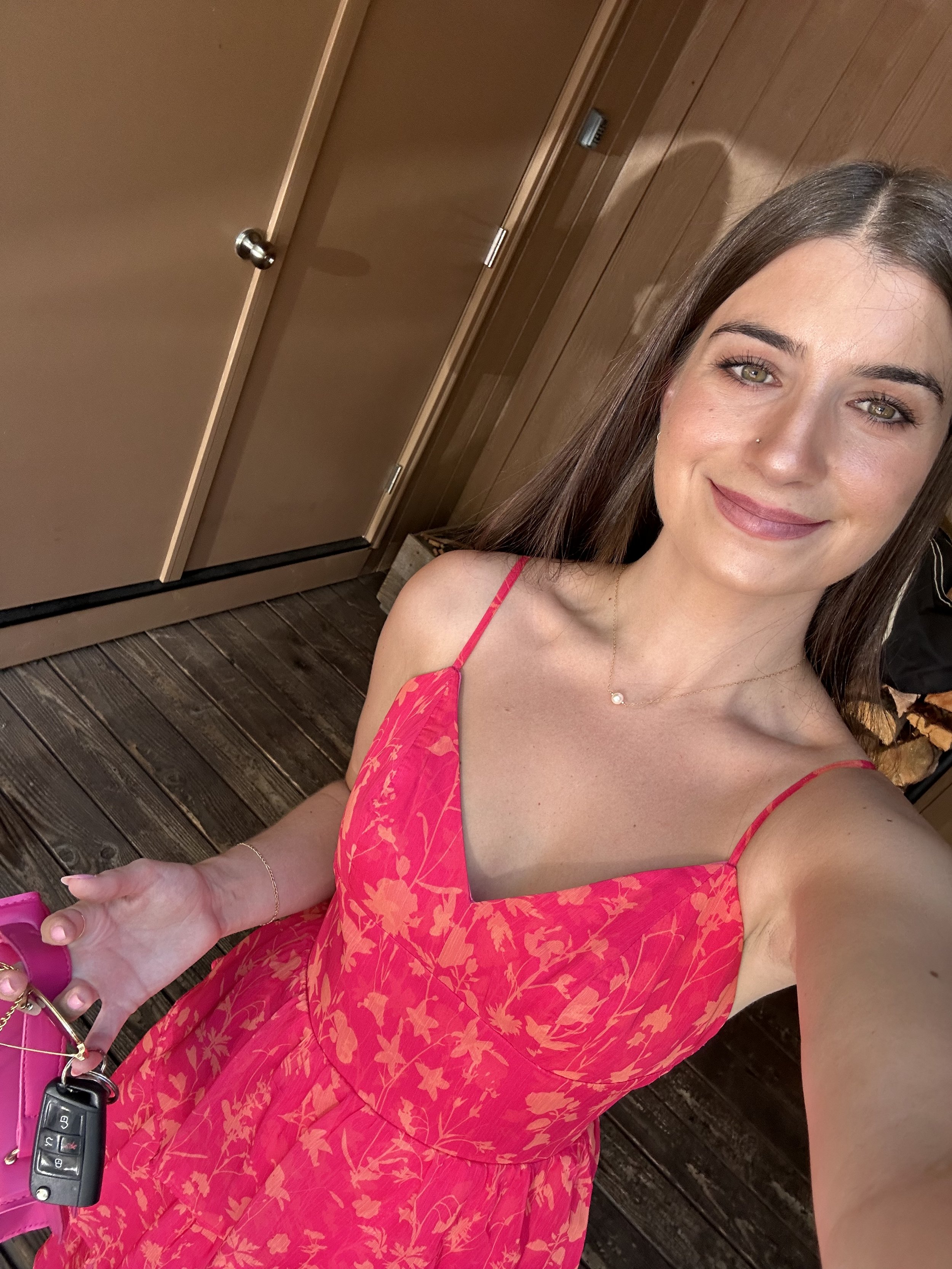 Young woman with long brown hair and light green eyes taking a selfie inside a wooden room, smiling, wearing a pink floral spaghetti strap dress, holding pink purse and car keys.