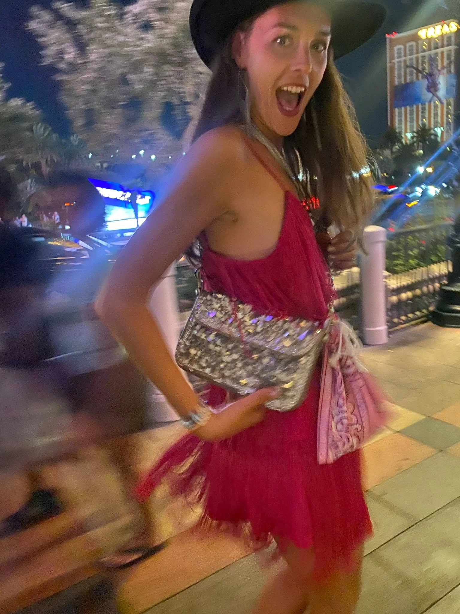 A woman wearing a red dress and a wide-brimmed black hat, carrying a silver, reflective clutch, smiling and posing on a lively city street at night with bright lights, palm trees, and an illuminated casino sign in the background.