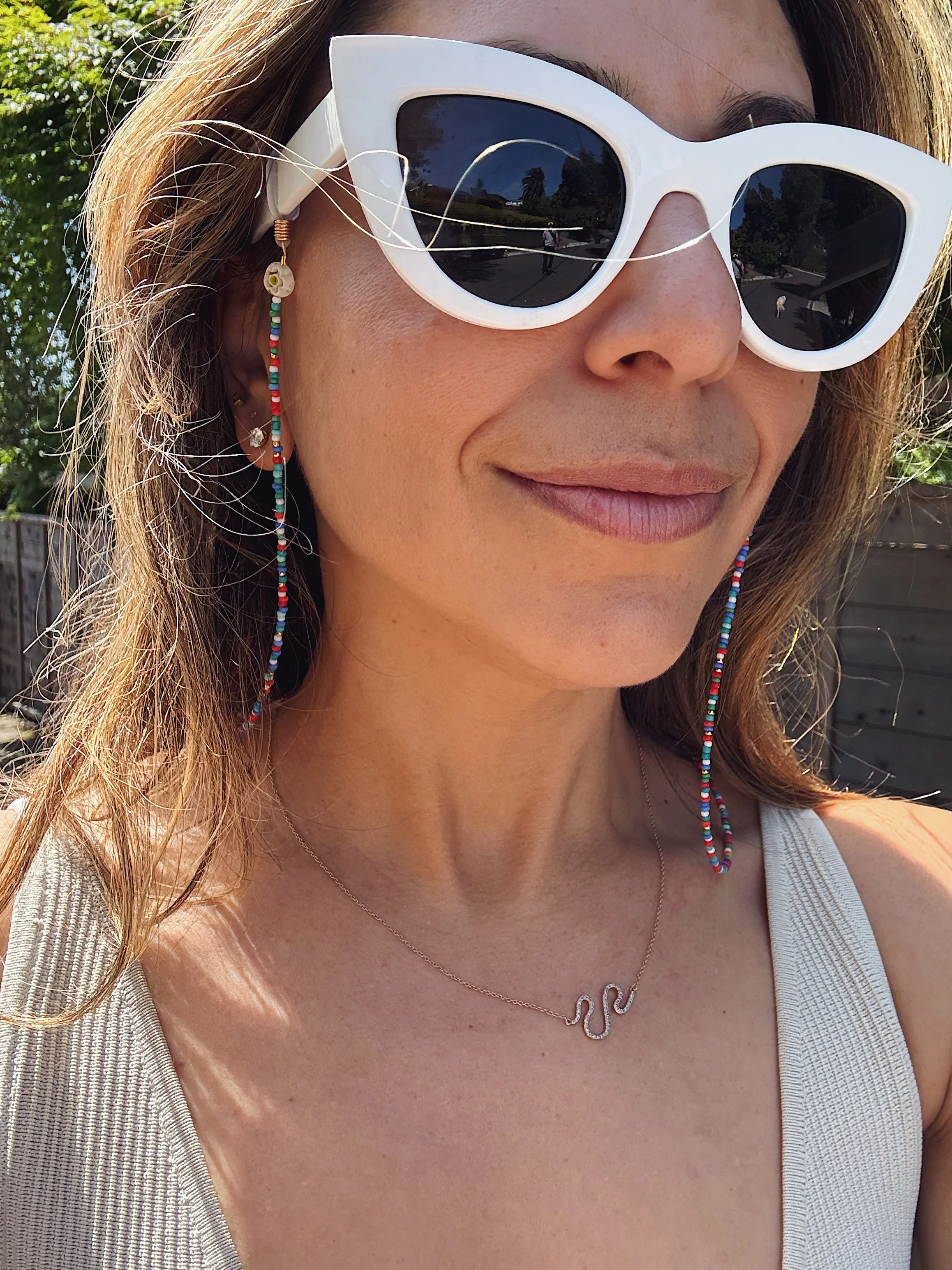 Close-up of a woman wearing white sunglasses, beaded earrings, a necklace with a snake pendant, and a sleeveless top outdoors on a sunny day.