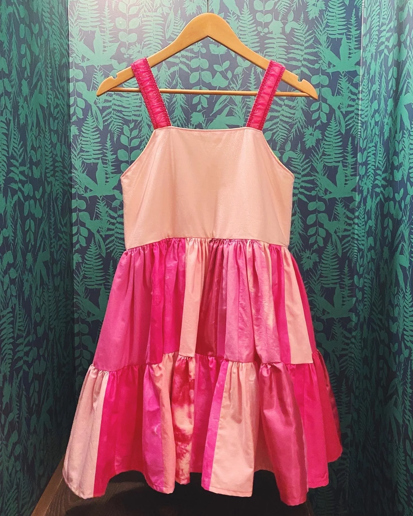 A pink and peach sleeveless dress with a tiered skirt hanging on a wooden hanger against a green leafy patterned wallpaper background.