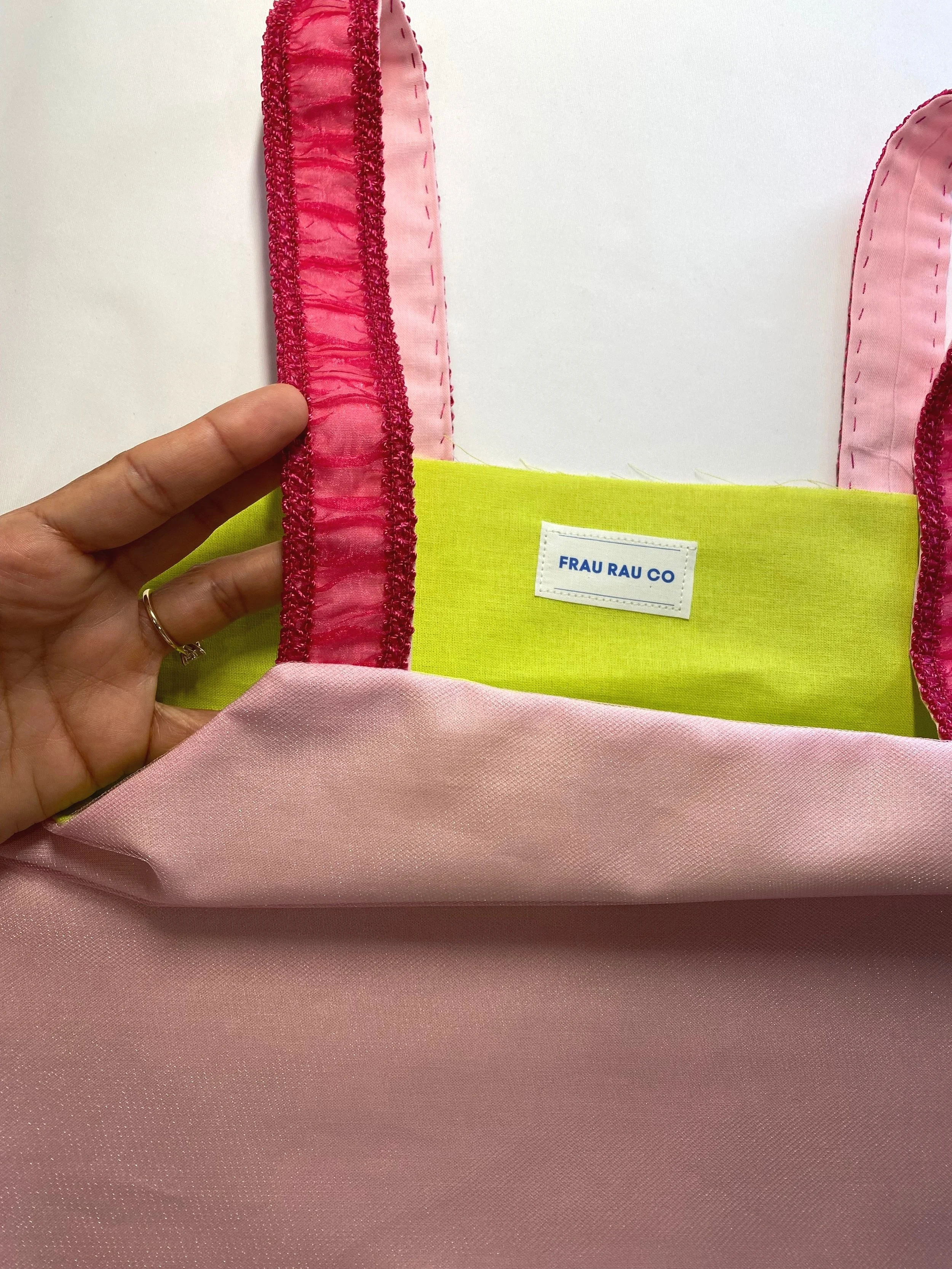 Close-up of a pink fabric bag with lime green and pink lining, held by a hand, showing a label that reads 'FRAU RAU CO' and pink straps with red decorative trim.