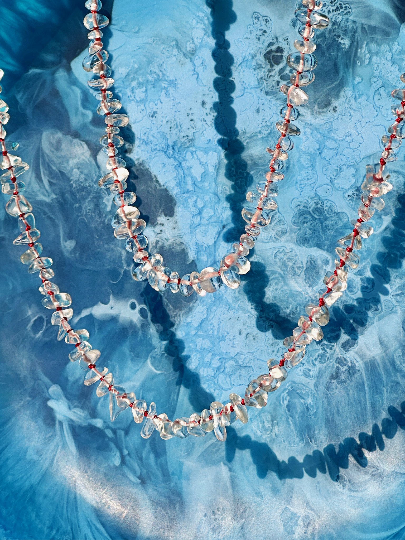 A clear bead necklace with red string, laid on a blue marbled surface with shadows cast by the necklace.