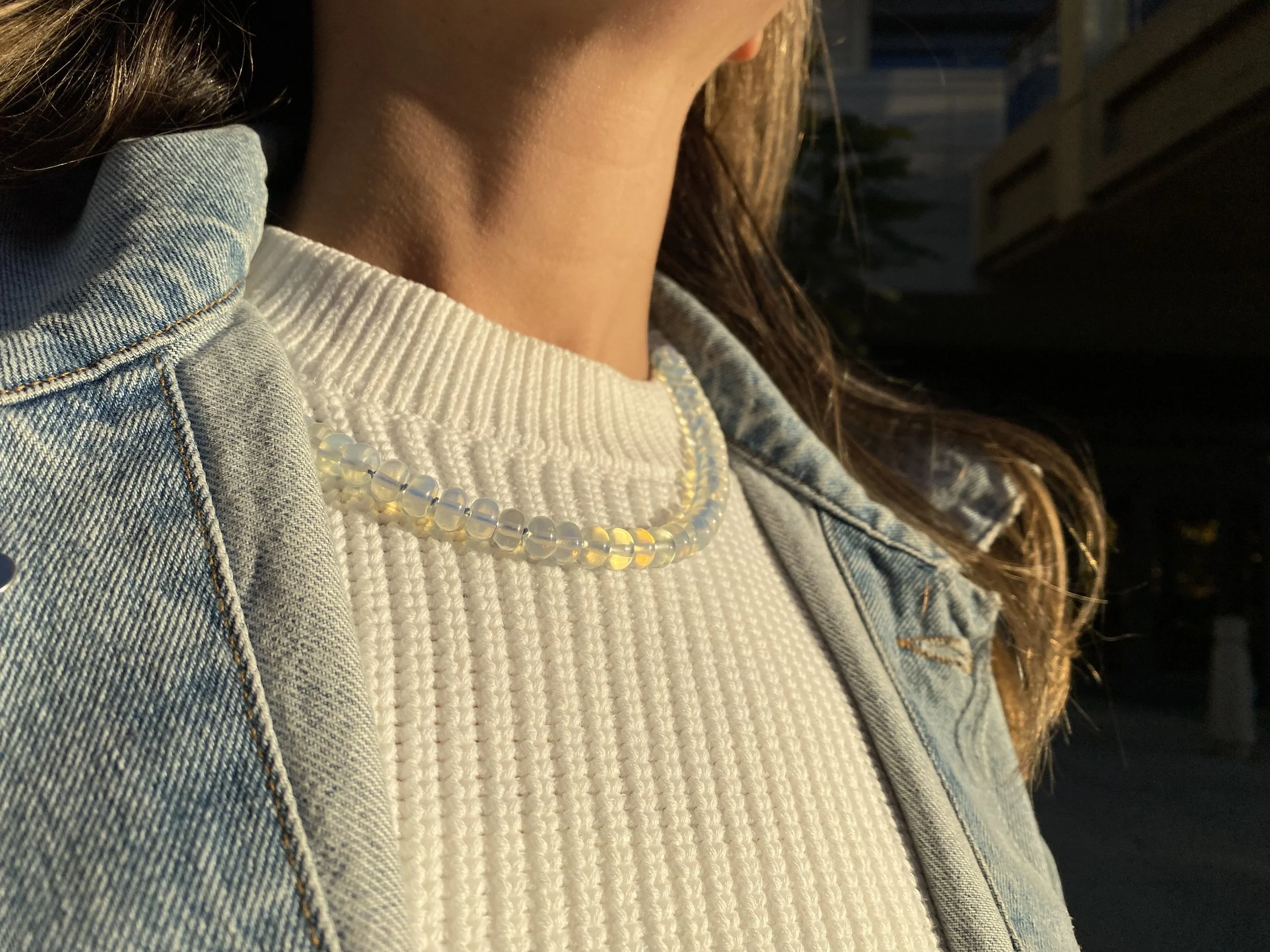 Close-up of a person's neck and chest, wearing a white ribbed sweater, a denim jacket, and a pearl necklace, with sunlight illuminating their skin and clothing.