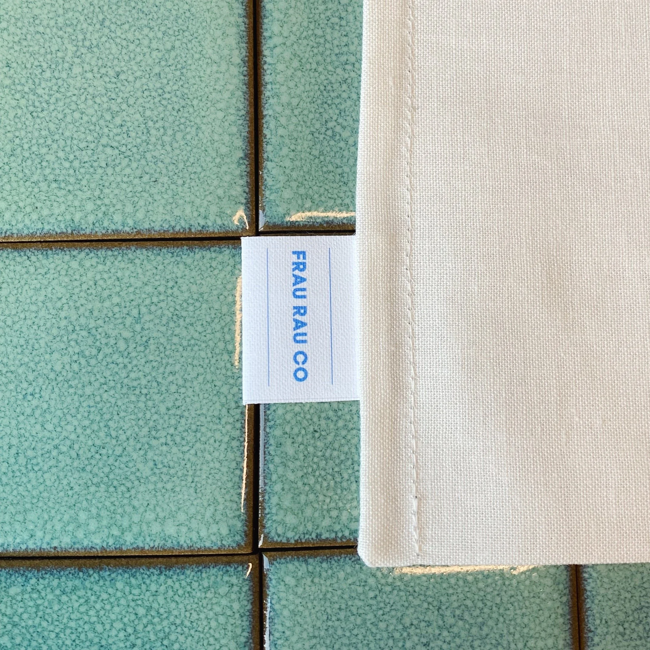 Close-up of green tiled surface with a white fabric piece and a label that reads 'FRAU RAU CO' in blue text, placed on the tiles.