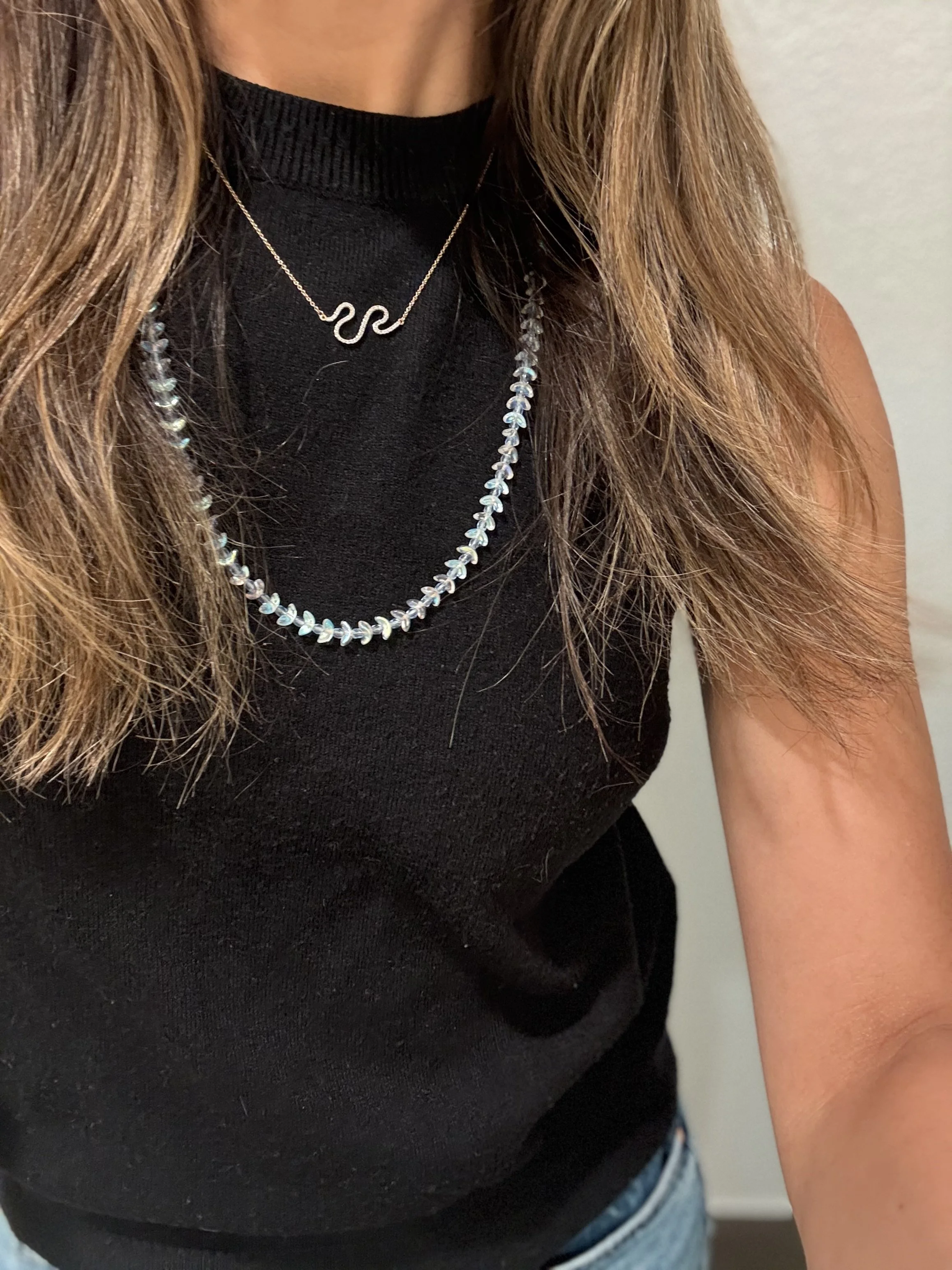 Close-up of a woman in a black sleeveless top, wearing two layered necklaces, one with a wave-shaped pendant and the other with small iridescent beads.
