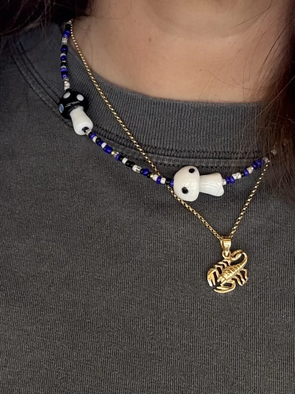 Person wearing two layered necklaces: one with white skull beads and the other with a gold scorpion pendant, over a gray shirt.