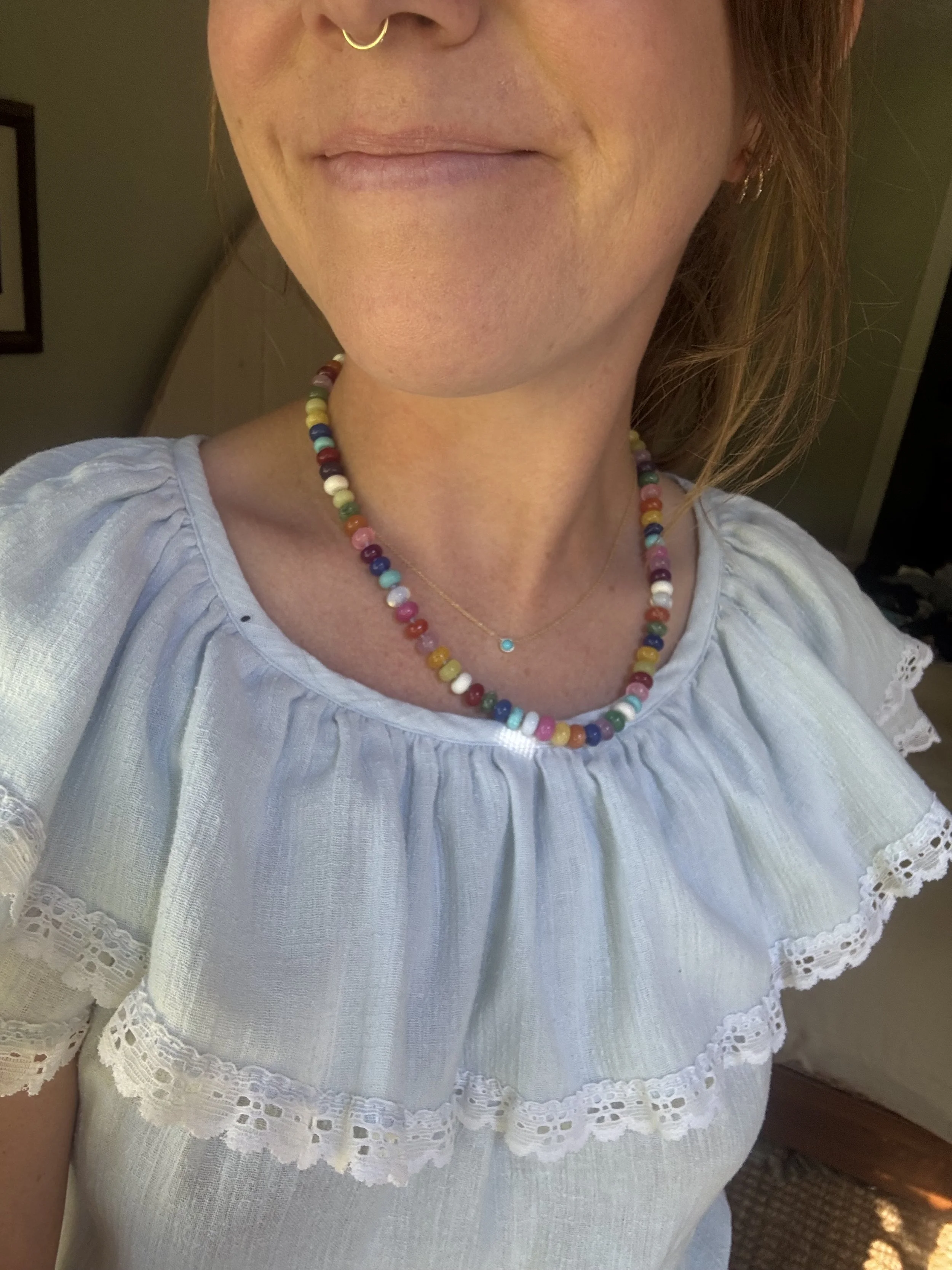 Close-up of a woman's neck and lower face, she is wearing a colorful beaded necklace, a delicate gold chain with a small turquoise pendant, a white embroidered blouse, and a gold nose ring.