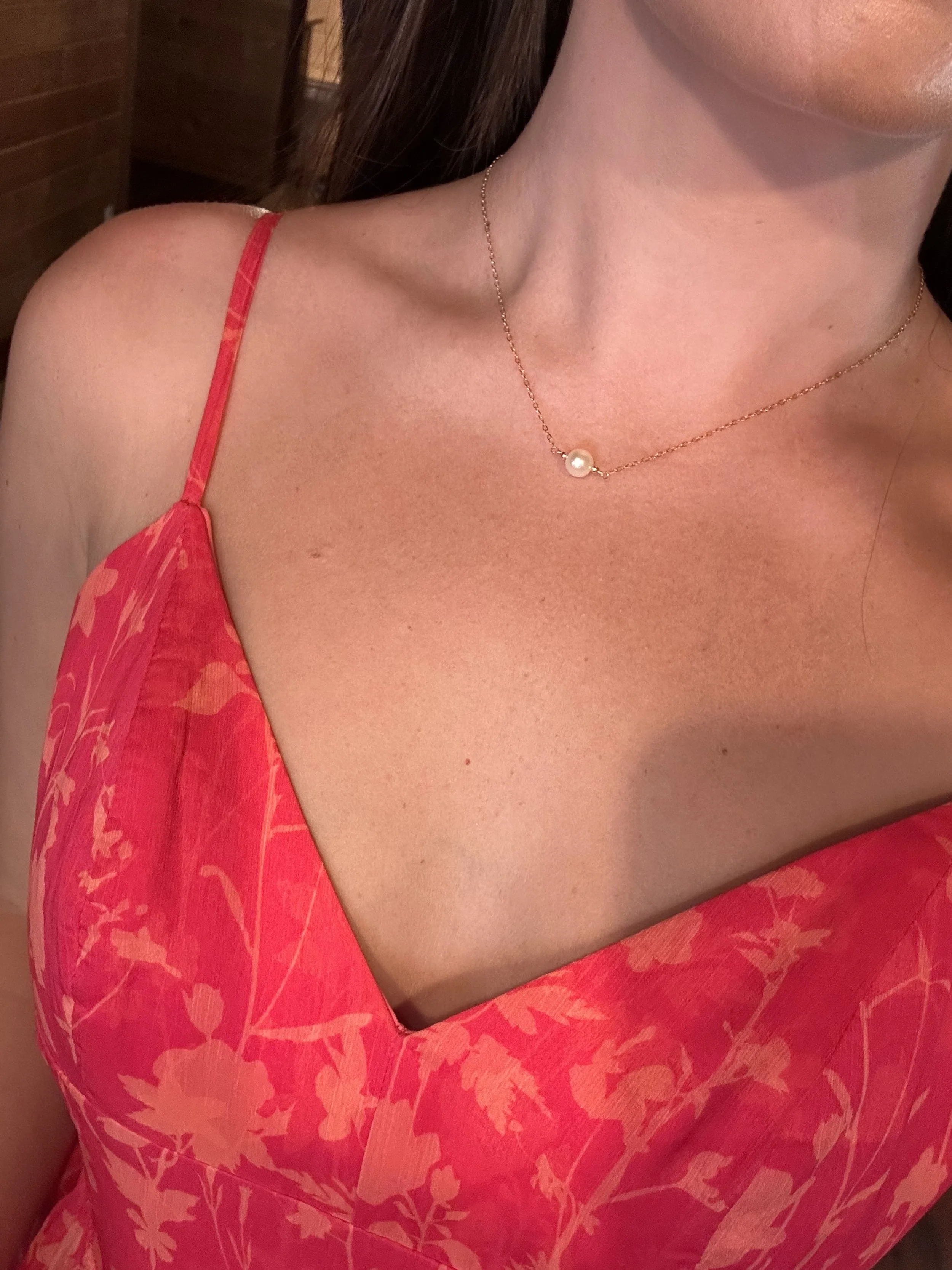 Close-up of a woman's upper body in a pink floral dress, showing her neck and collarbone, with a delicate gold necklace featuring a single pearl pendant.