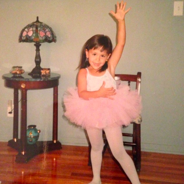 A young girl dressed as a ballerina wearing a pink tutu, white tights, and a white top, standing in a room with hardwood floors and a light blue wall. She is smiling, with one arm raised and the other hand on her chest, and is positioned near a wooden chair. Behind her is a side table topped with a decorative lamp and some miscellaneous items.