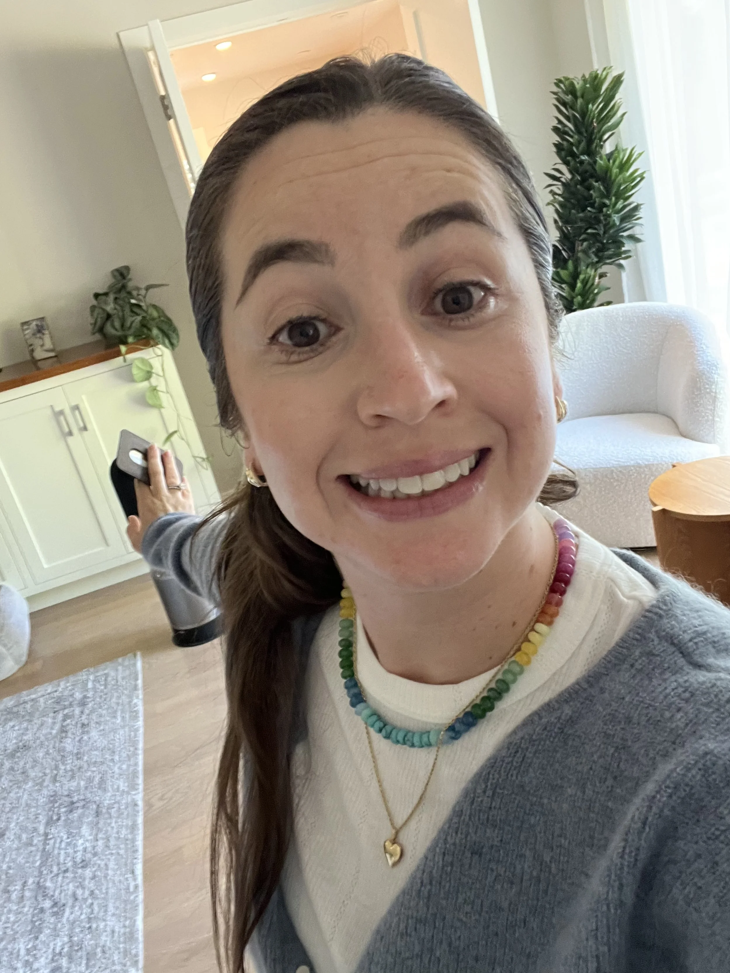 A woman with brown hair and fair skin smiling at the camera, wearing a colorful beaded necklace, a white top, and a gray cardigan. She appears to be in a cozy living room with a white armchair, a wooden coffee table, and some plants in the background