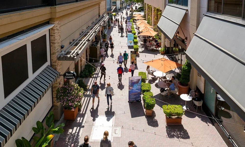 Pedestrian street with outdoor seating, umbrellas, potted plants, and shops with awnings.