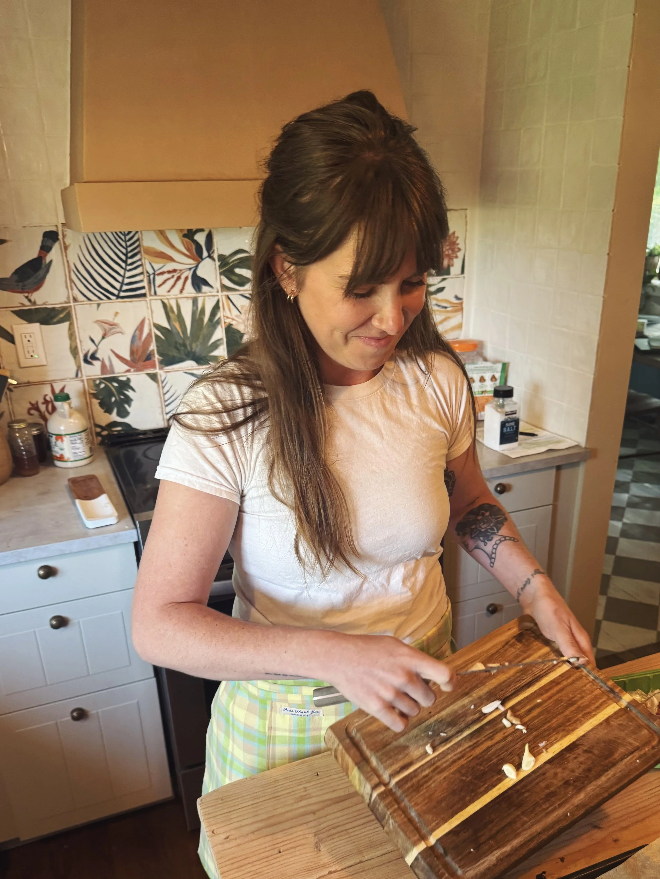 A woman in a white t-shirt and yellow checkered apron is chopping garlic on a wooden cutting board in a kitchen.