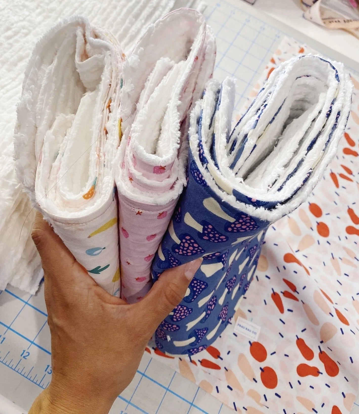 A hand holding several rolled-up cloth diapers with colorful patterns, in front of a blue and white patterned fabric and some orange and pink fabric pieces on a cutting mat.