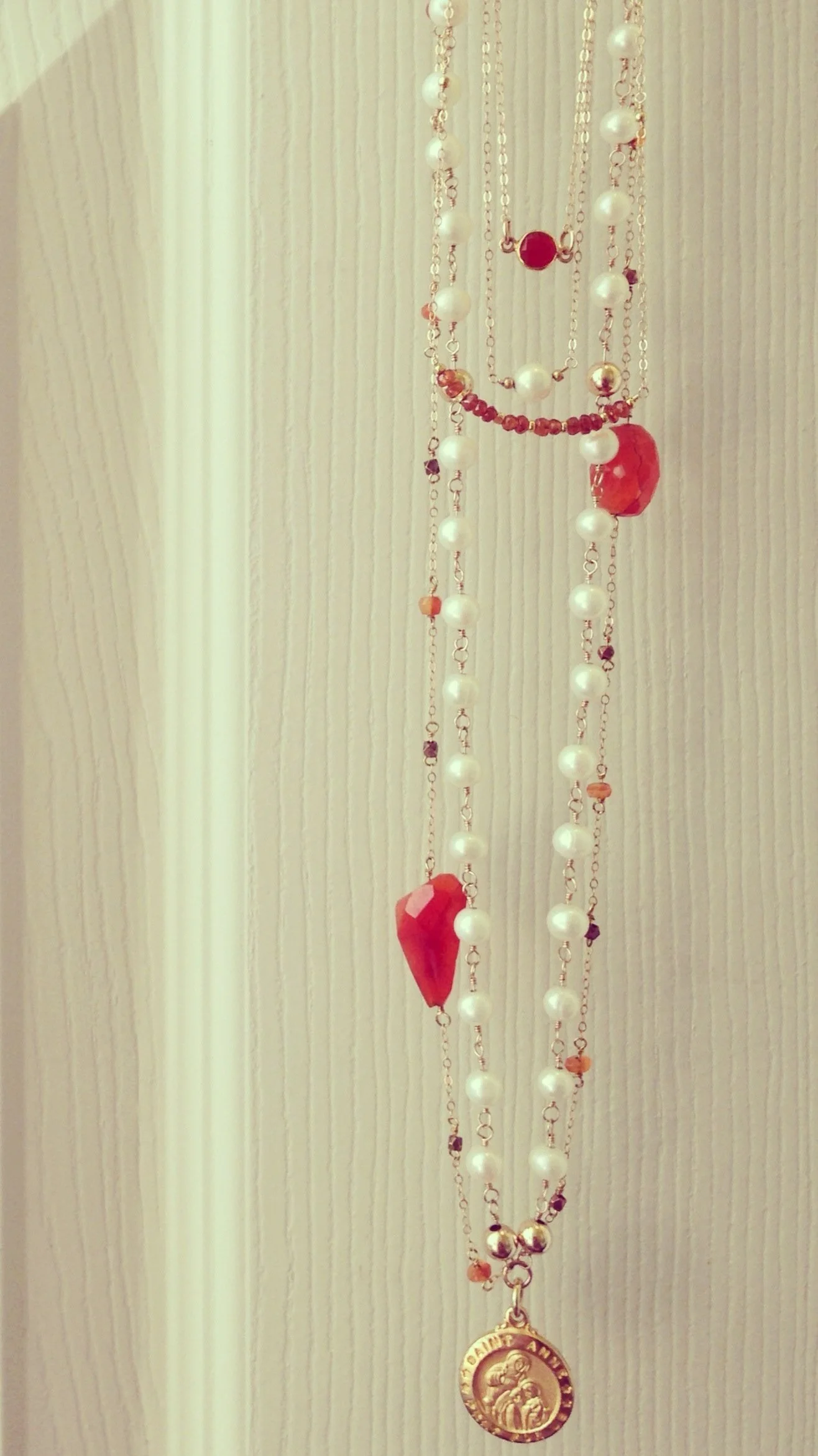 Close-up of layered necklaces hanging against a white textured wall, featuring pearls, red beads, a large red heart-shaped pendant, and a gold religious medal.