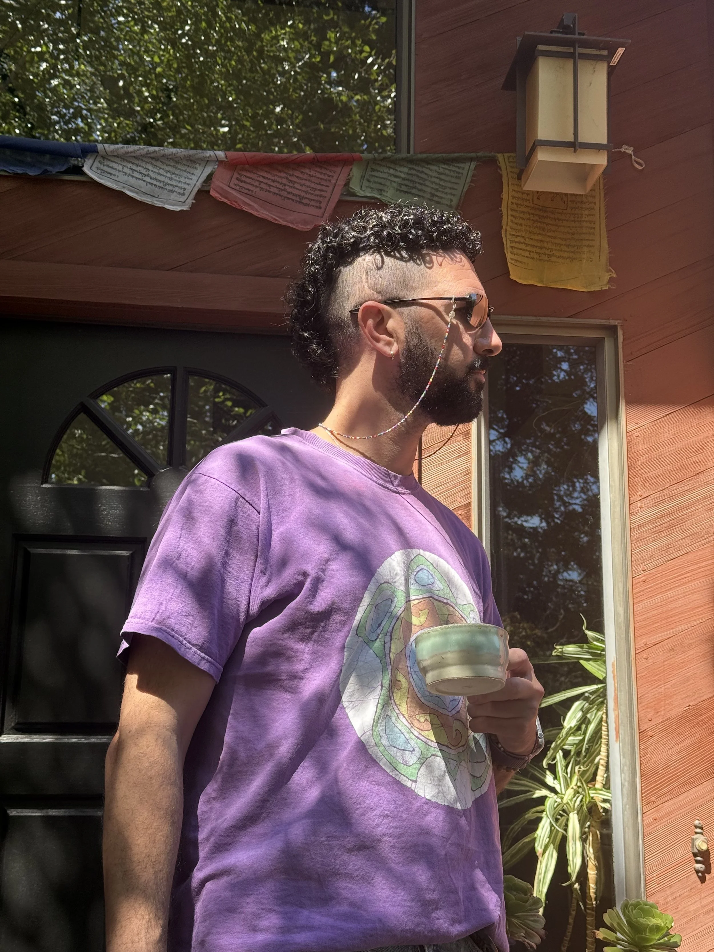 A man wearing a purple T-shirt with a colorful graphic, sunglasses with a chain, and holding a ceramic mug while standing outside near a building with a window and plants.