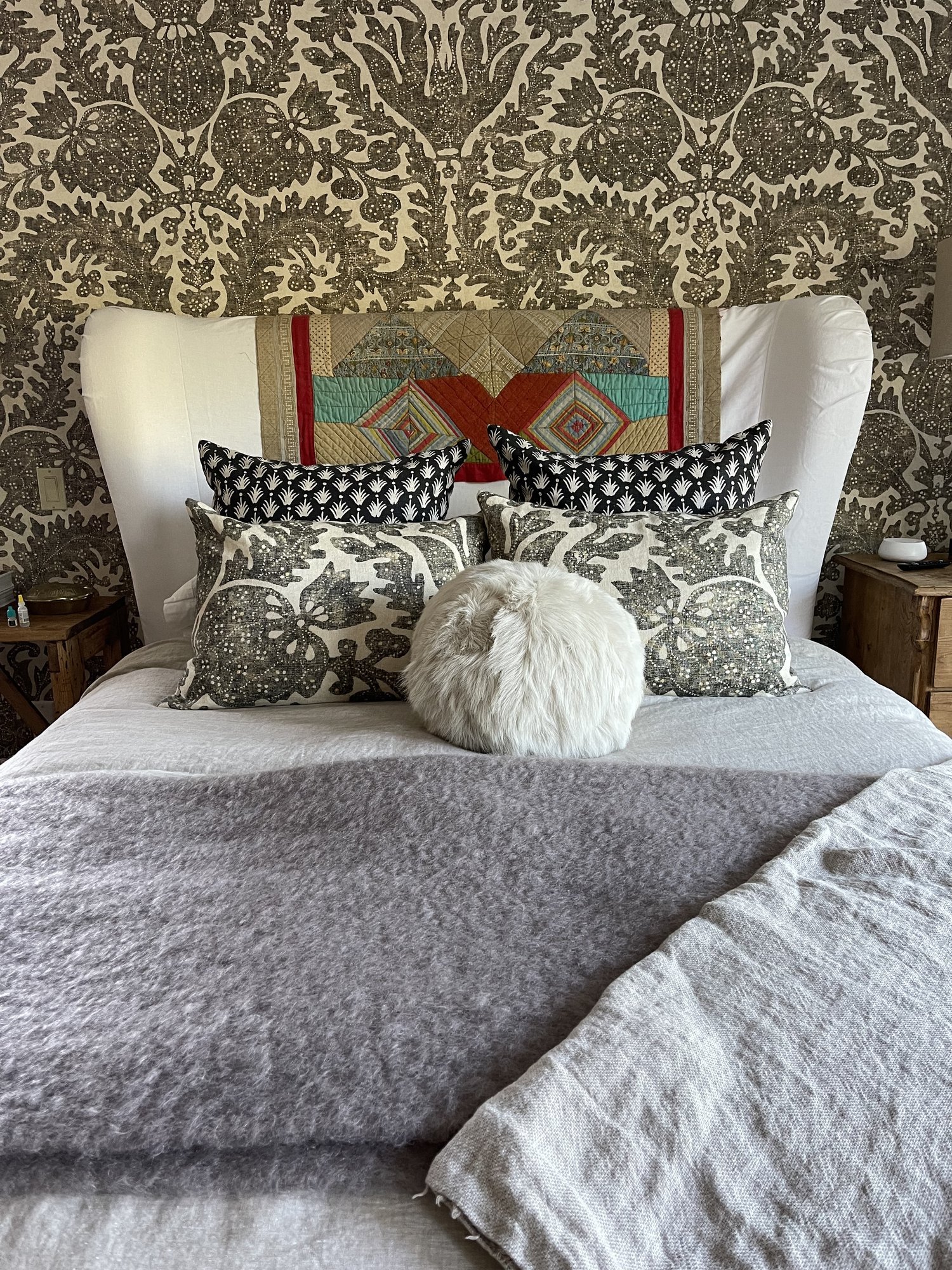  A bed with a flared white headboard with a quilt laid over it. The bed has four black and white patterned pillows and one fluffy round white one arranged neatly. The wall behind the bed is wallpapered in large print green abstract florals.  