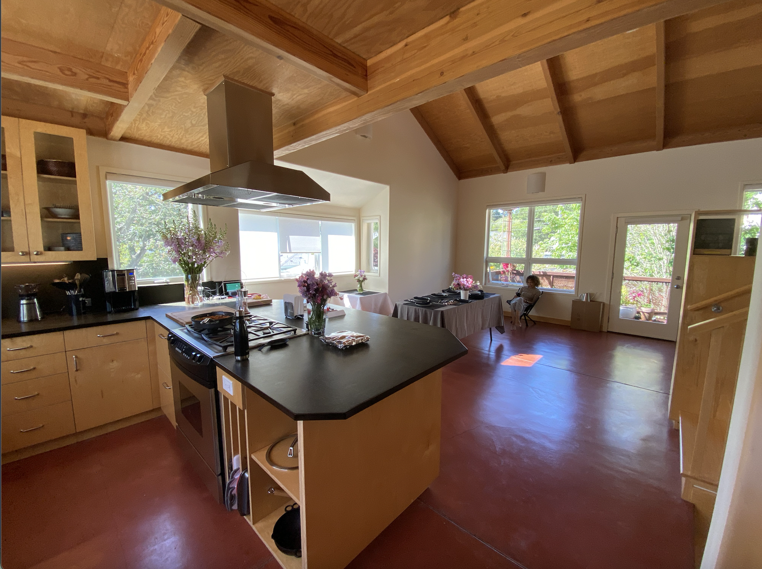  An open kitchen and living space. The space is separated by a countertop with black surface, oven, and wooden cabinets. There is a hanging range vent, red concrete floors, and large windows on two walls.  