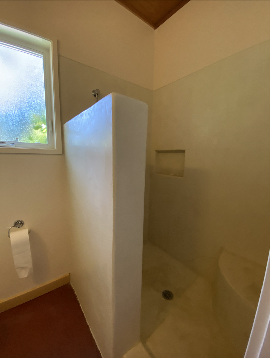  A bathroom with off-white walls and a half wall enclosing a shower. There is a window above a toilet paper holder on the wall.  