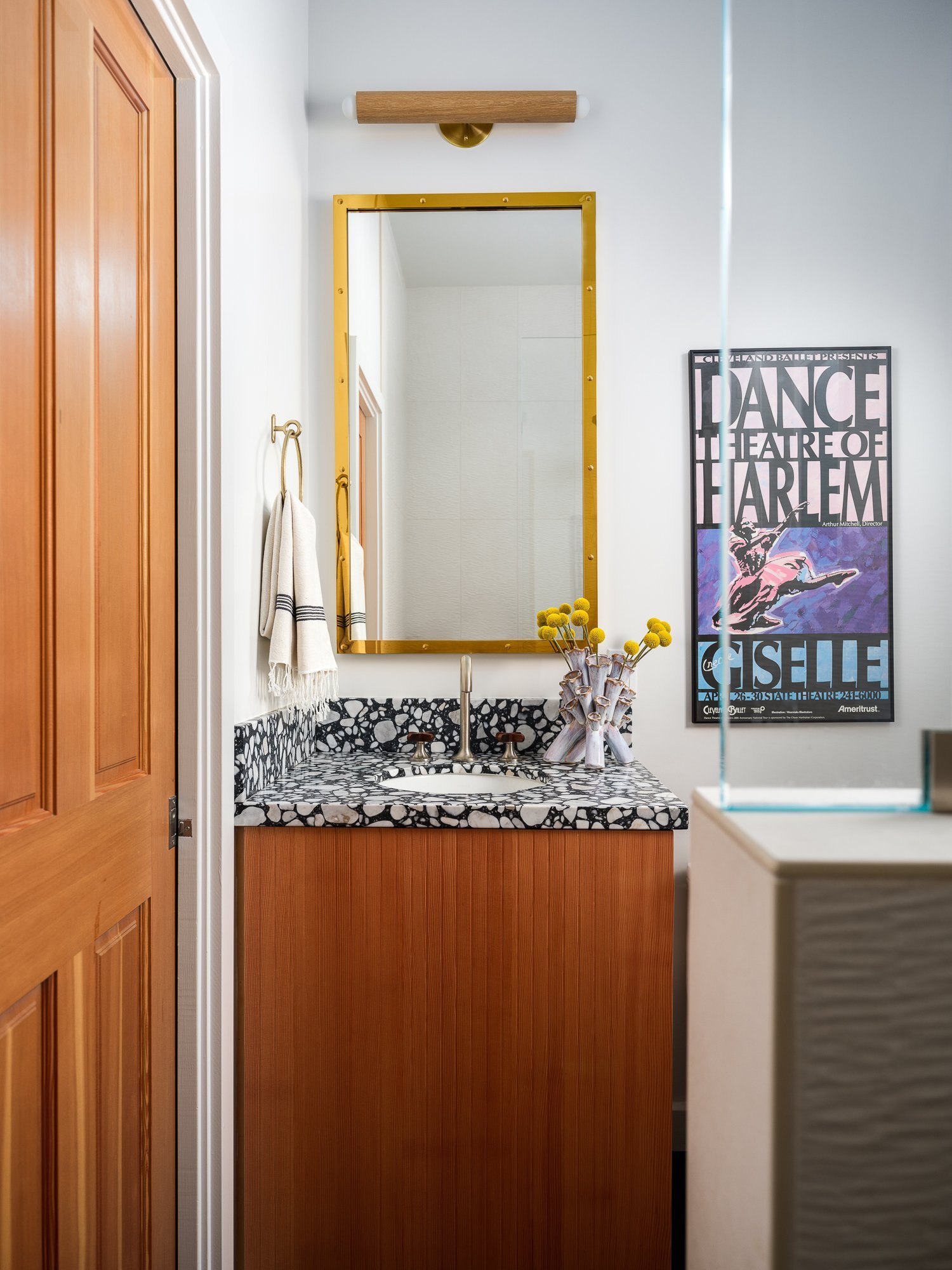  A bathroom with a wooden door and half-wall with glass top enclosing the shower. The vanity cabinet is wood with black and white terrazzo tile countertop and a small circular sink. A gold framed mirror hangs above the sink next to a framed vintage “