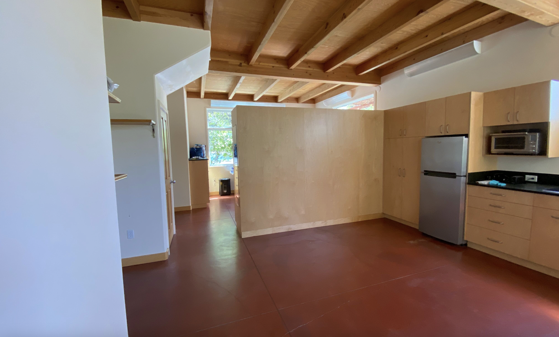  Open kitchen with red concrete floor, light wood ceiling, light wooden cabinets, and a silver refrigerator. There is a wooden partition wall separating the kitchen from an adjoining room with window.  