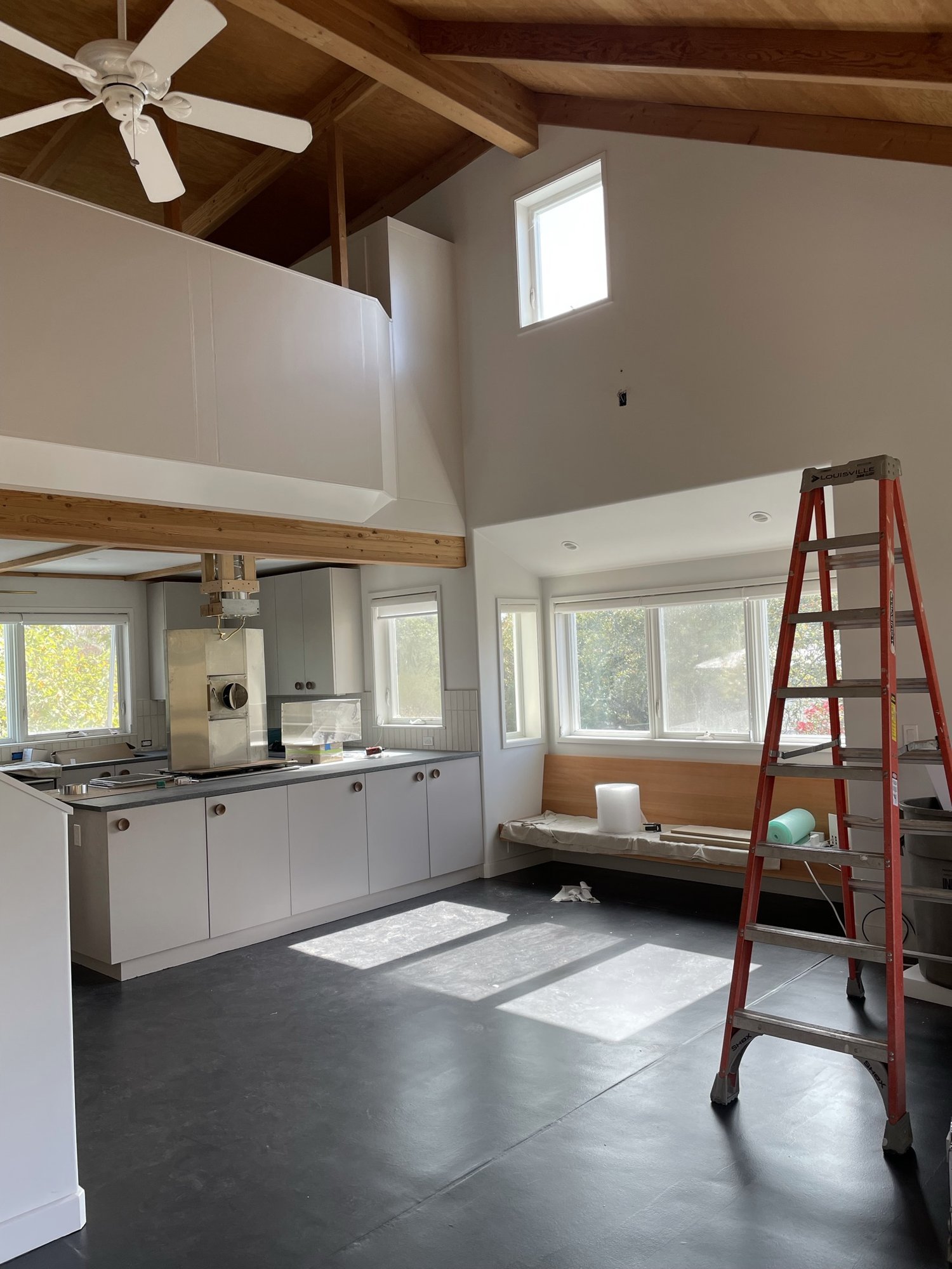  A progress image of a living space under construction. There is a ladder in one corner and a built-in bench under construction below a window. The kitchen cabinet extends to separate the living from kitchen space and has white cabinets with large ci