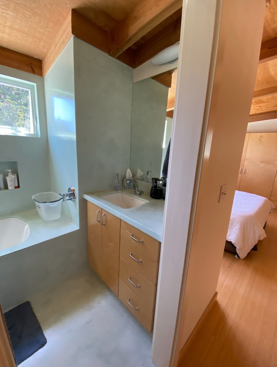  A bathroom with a small vanity with a light countertop and wooden cabinetry. The bedroom is visible past the open door.  