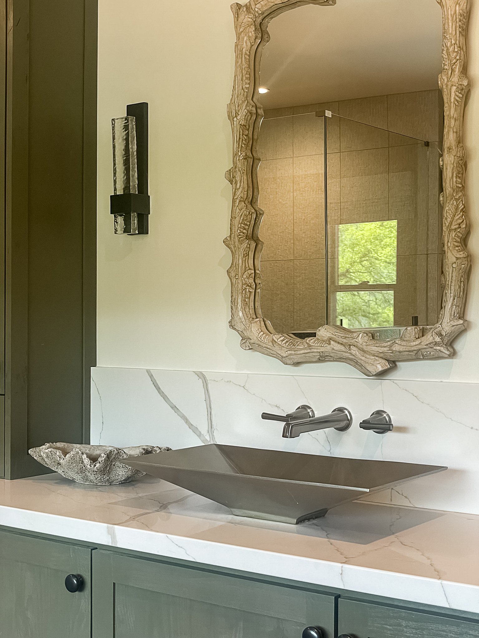  A bathroom with a wood framed mirror, marble countertop, square bowl style sink, seashell soap holder, and green cabinets. 