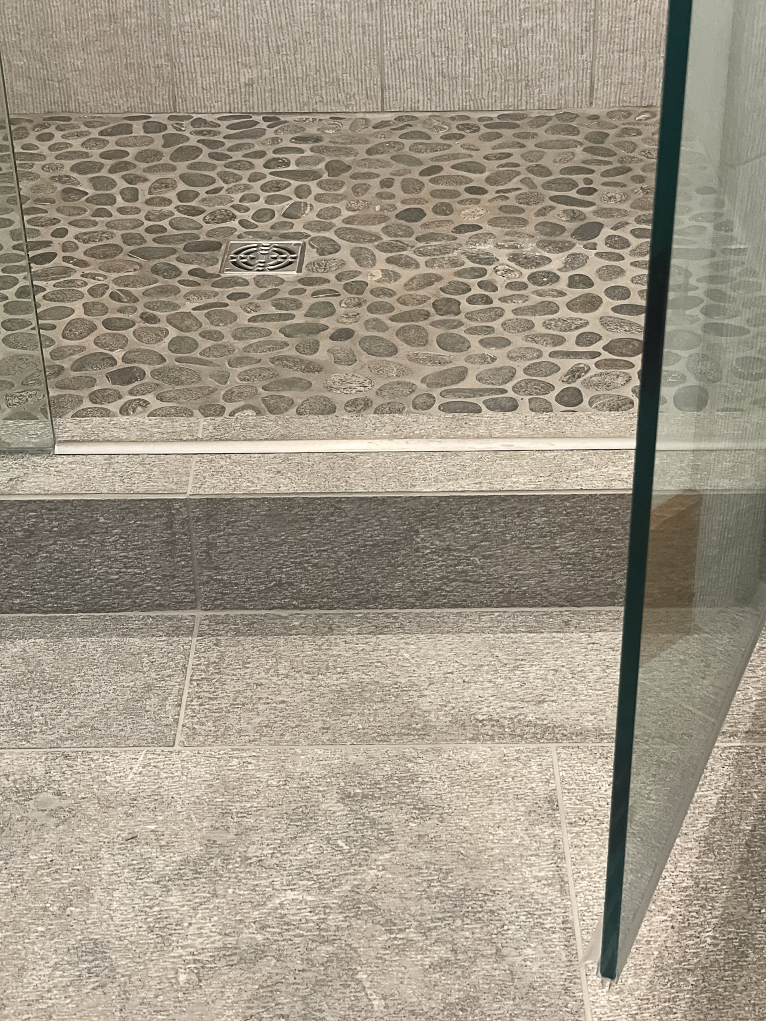  A bathroom floor with small grey river stone shower floor next to a textured grey stone tile floor.  