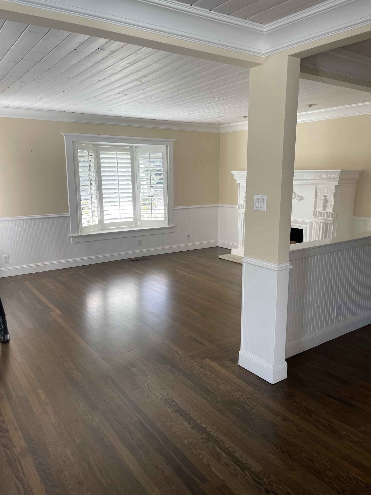  Before image of a low ceilinged living space with wood floors, pale yellow walls, and painted white ceilings. There is a column and half wall separating the space and a large white fireplace along one wall.  