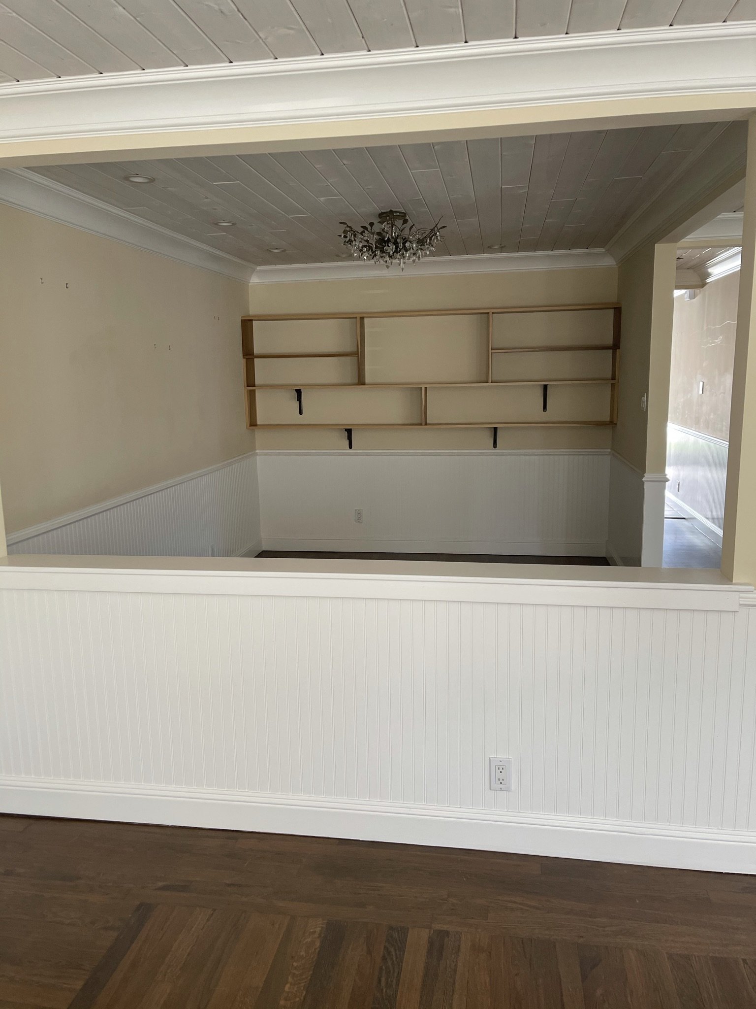  A before image of a living space separated by a low half wall. Beyond the half wall is a small space with a shelving unit on the wall.  