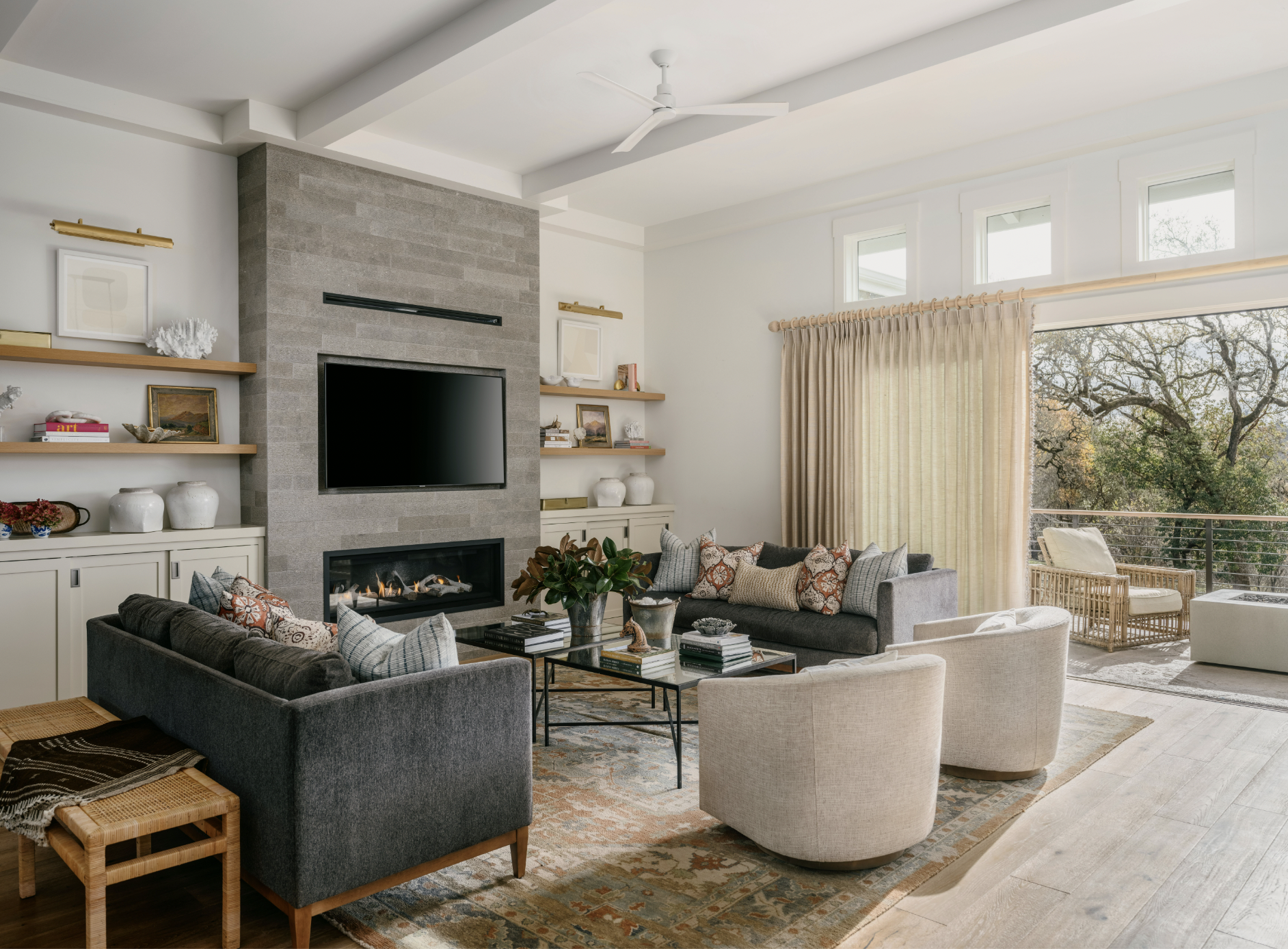 A living area with a modern stone fireplace. A TV is inset above the fireplace. Chairs and two couches surround a coffee table at the center of the room. Floating wooden shelves hold curated objects on both sides of the fireplace.  