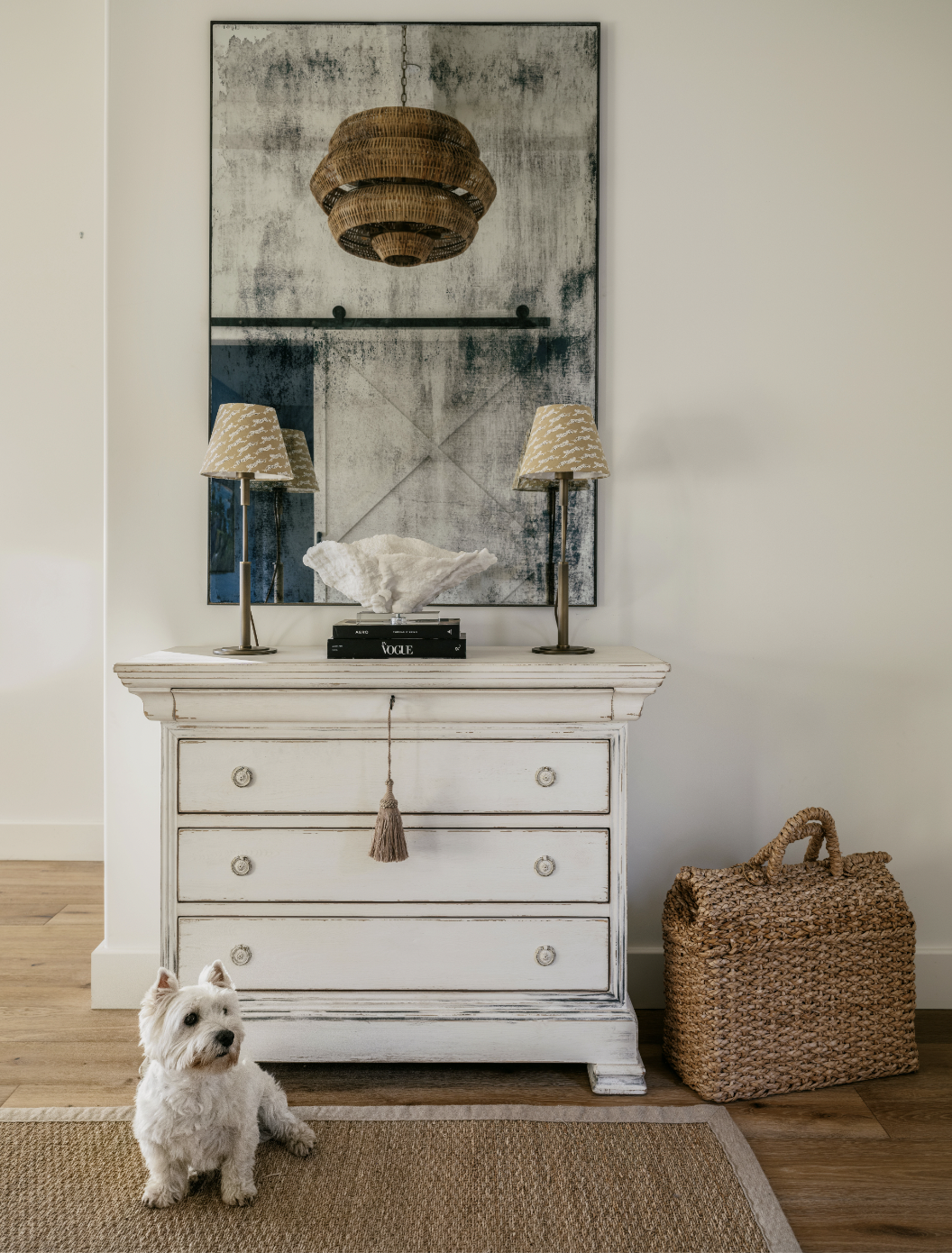  An antique white dresser with a large art piece hung above it. A small white Westie sits in front of the dresser.  