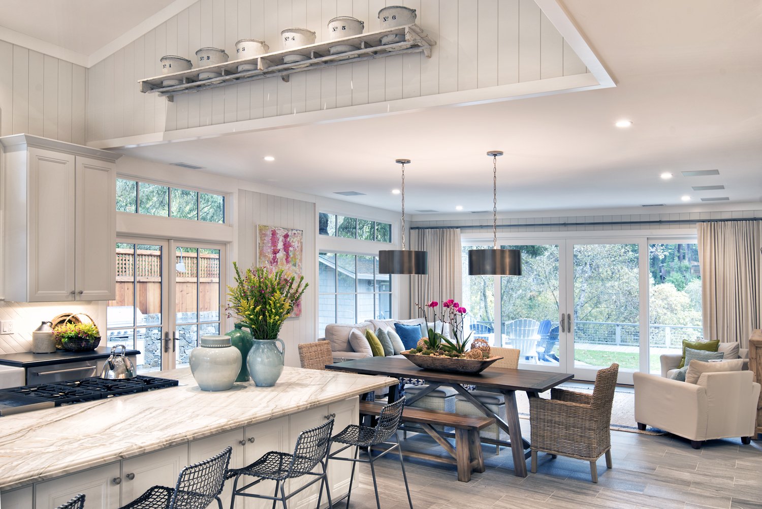  A living space with a kitchen island, dining table, and couch all visible. The room is surrounded by large, windows to the backyard.  