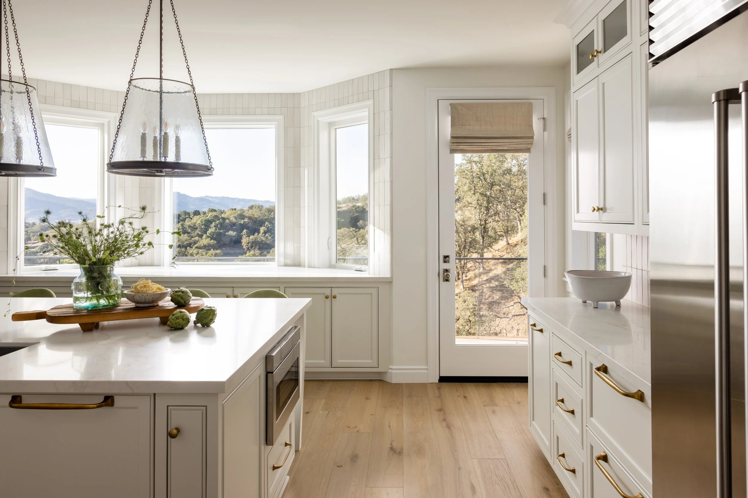 Bright white kitchen with panoramic hillside views, quartz island, brass hardware, and seeded glass pendant — Bartlett Residence by Tama Bell Design, Ukiah CA