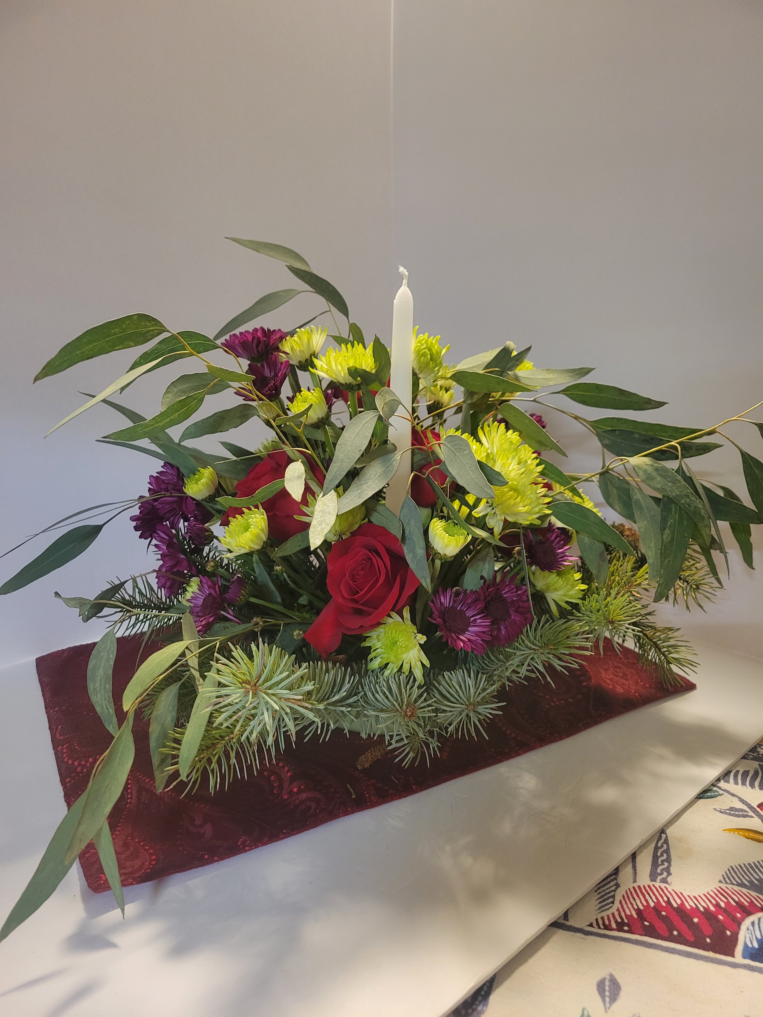 A floral arrangement with red roses, yellow and purple chrysanthemums, green leaves, and pine branches, placed on a red cloth atop a white table.