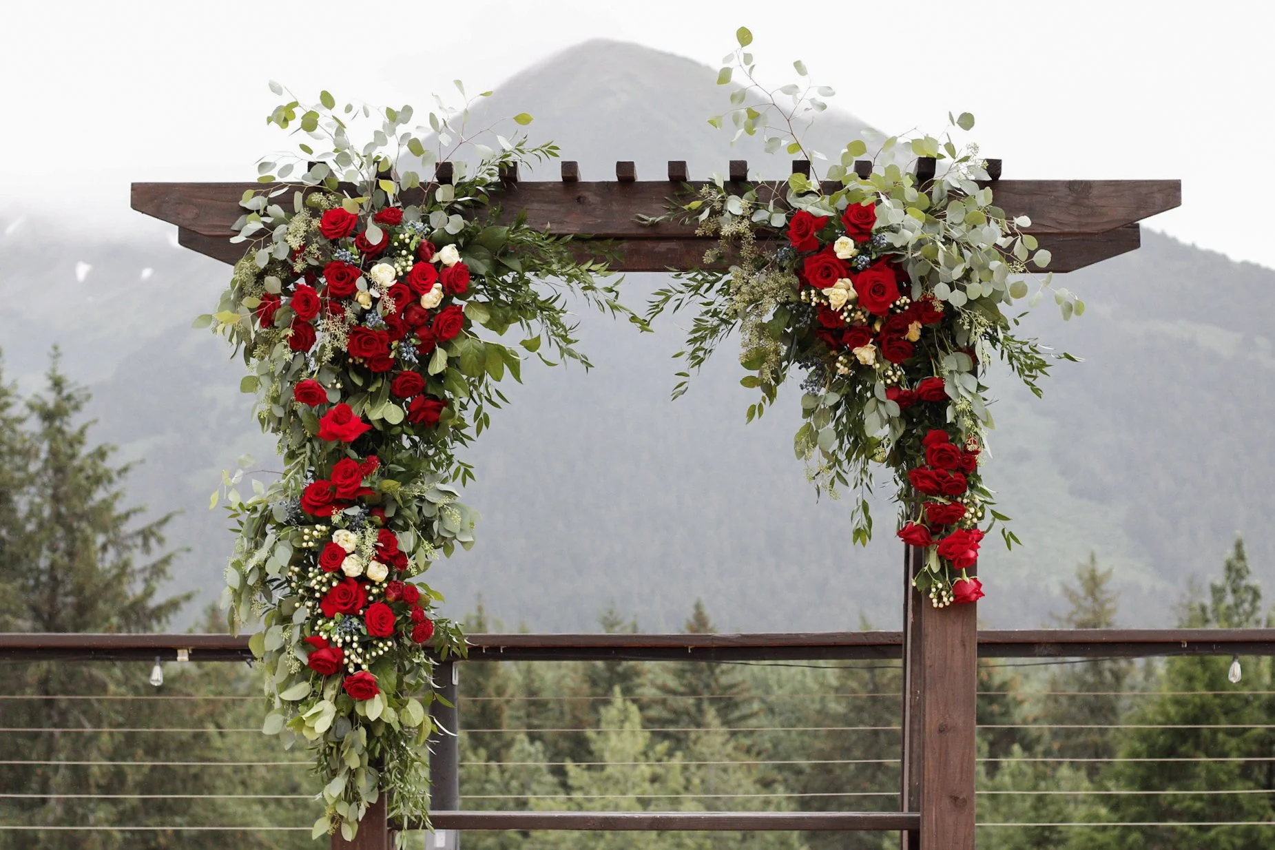 Wooden wedding arch decorated with red and white roses, eucalyptus leaves, and other greenery, set outdoors with mountains in the background.