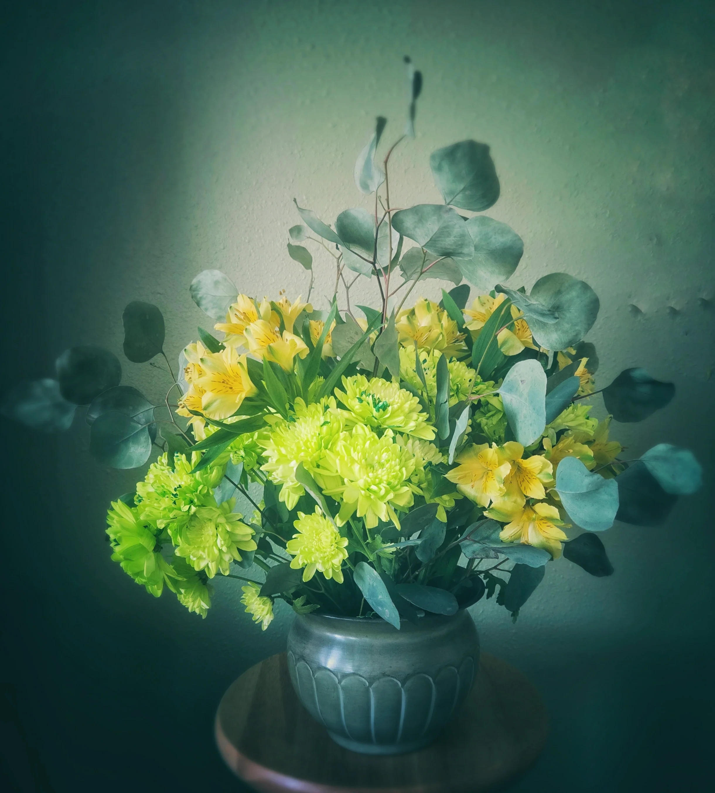 A bouquet of yellow and green flowers with leafy greenery in a silver vase on a dark surface against a textured light green background.