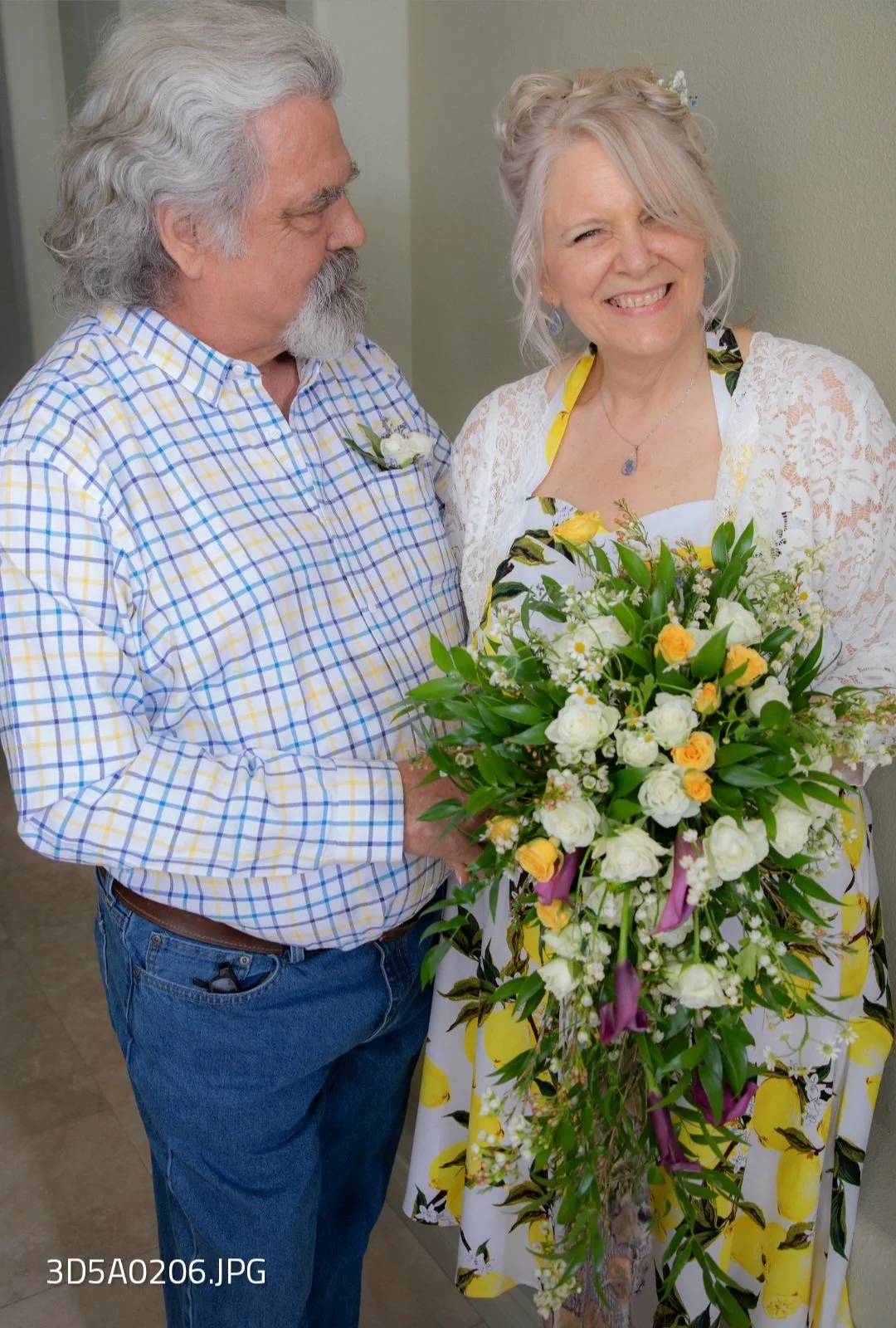 An older woman holding a large bouquet of white and yellow flowers, smiling and standing next to a man, both dressed in casual clothing, inside a room with beige walls.