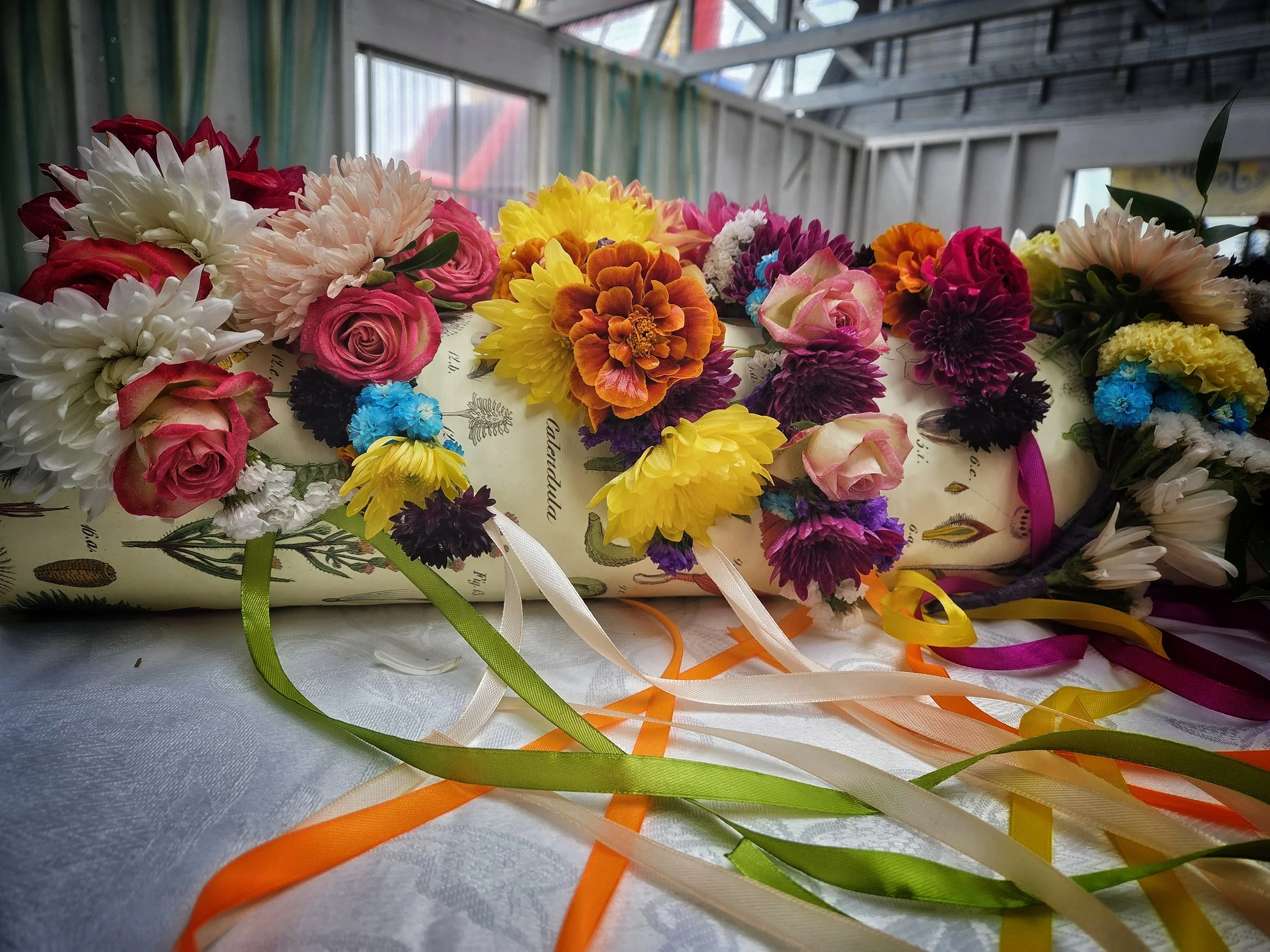 A display of colorful artificial flowers, including pink, yellow, and white blossoms, arranged on a surface outdoors near a busy road with cars, trees, and buildings in the background.