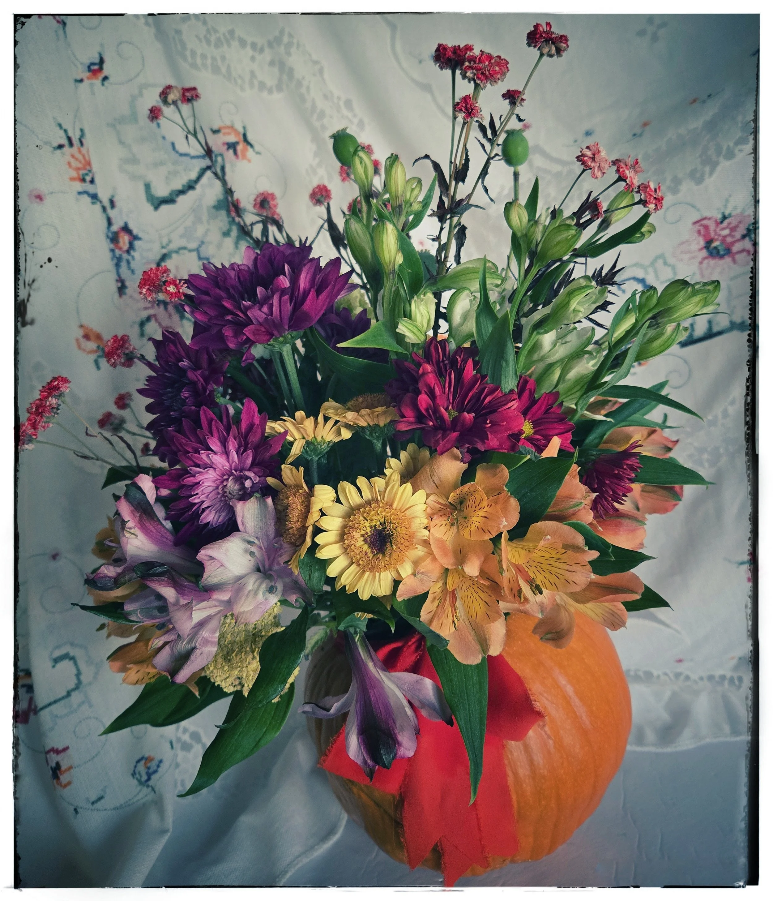 A bouquet of colorful flowers including purple, pink, yellow, and orange blooms arranged in a vase placed on an orange pumpkin, with a white lace tablecloth and embroidered cloth in the background.