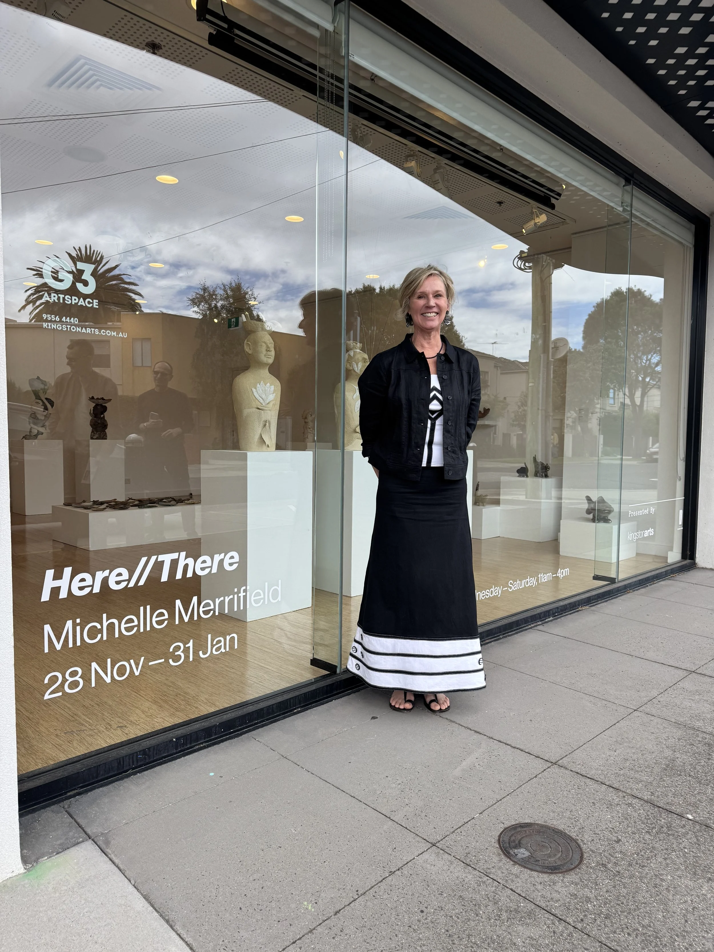A woman with short blonde hair, wearing a black jacket and a long black skirt with white stripes at the bottom, stands outside an art gallery window, smiling. The gallery sign reads 'Michelle Merrifield' and indicates an exhibition from November 28 to January 31. Inside the gallery, art sculptures and artworks are displayed.