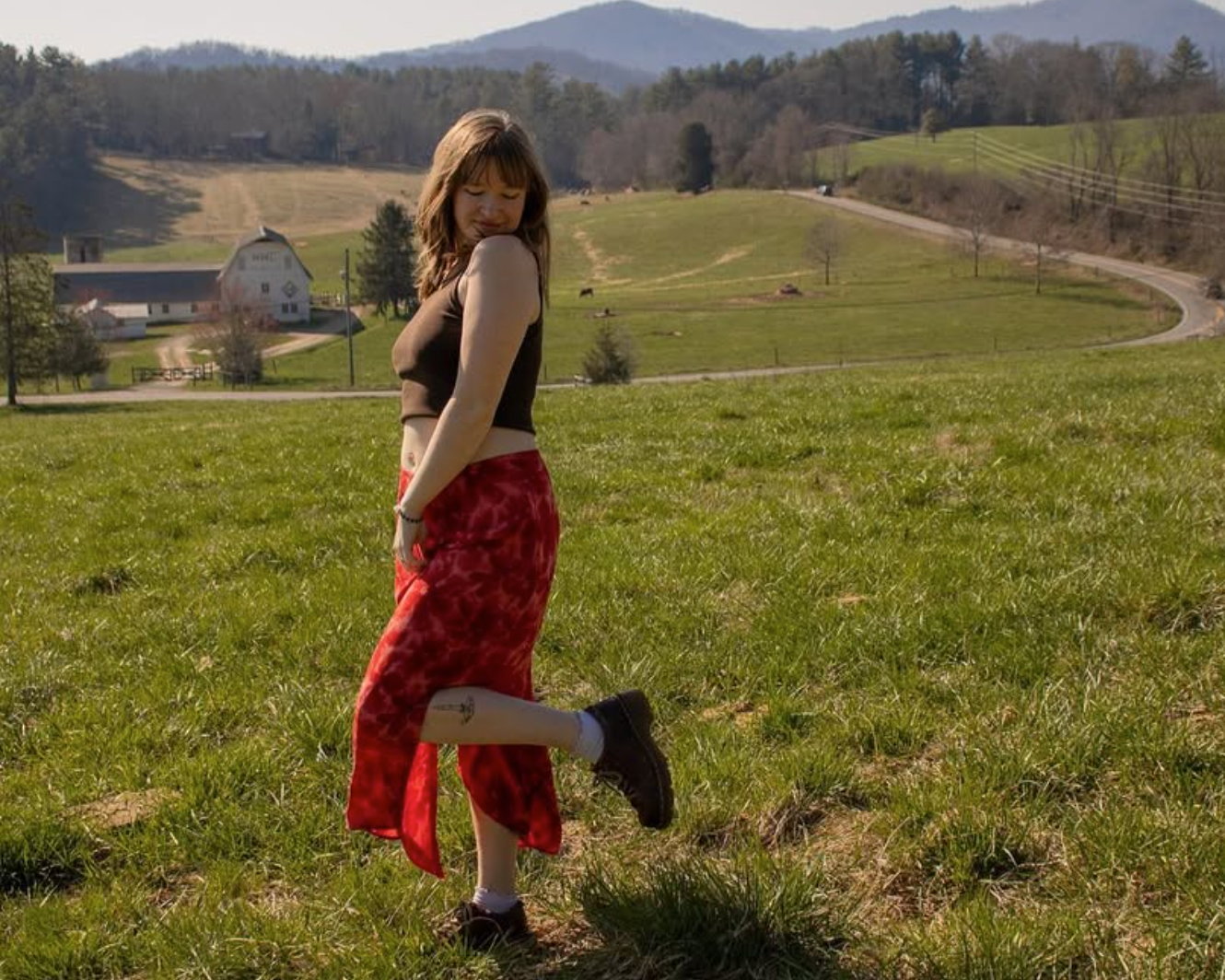 A girl in a skirt smiling in an open field, with a barn and mountains in the background
