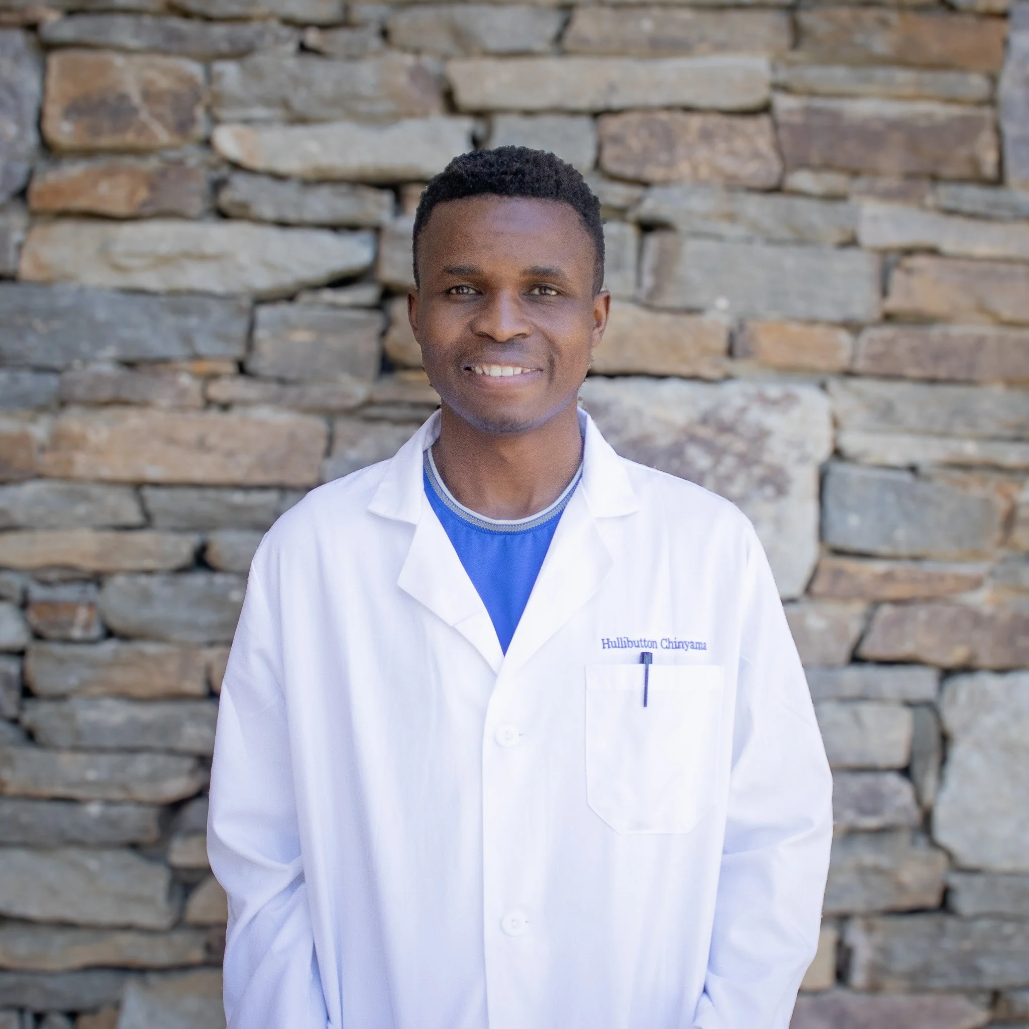 A young man smiling in a lab coat, with a stone backdrop